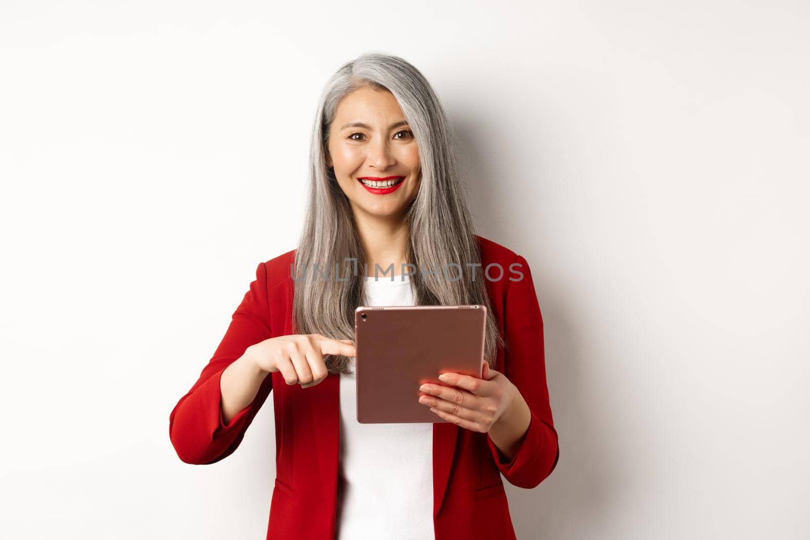 Business. Smiling senior businesswoman using digital tablet, pointing finger at screen, standing over white background by Benzoix