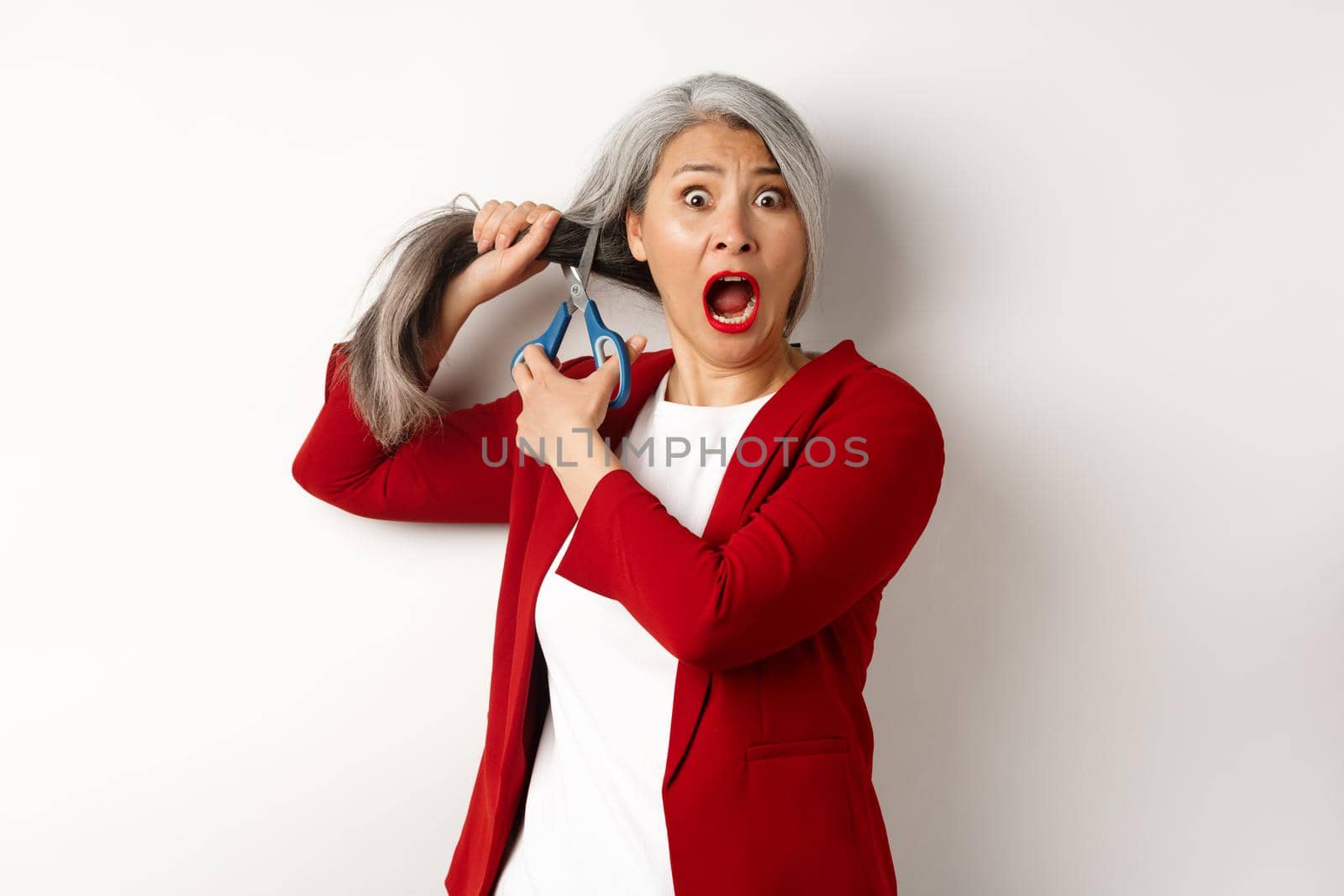 Scared asian senior woman cutting hair with scissors and screaming in panic, standing over white background.