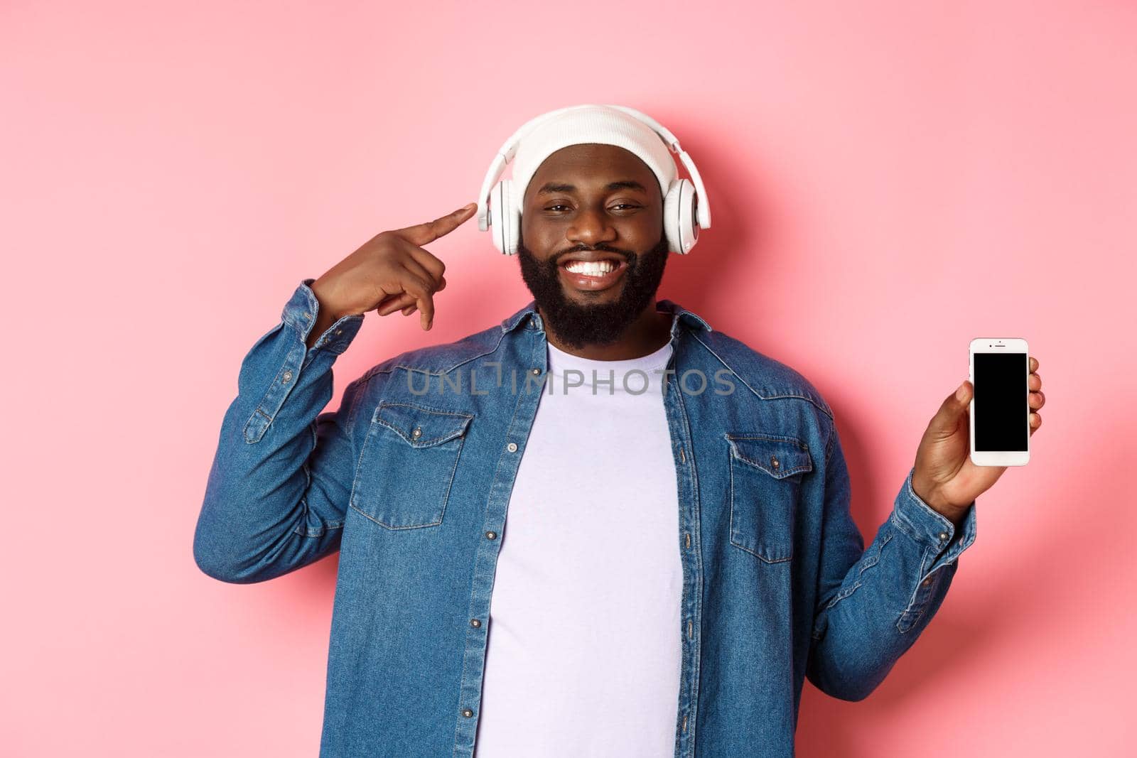 Happy black man listening music, pointing finger at headphones and smiling, showing mobile phone screen app or playlist, standing over pink background.