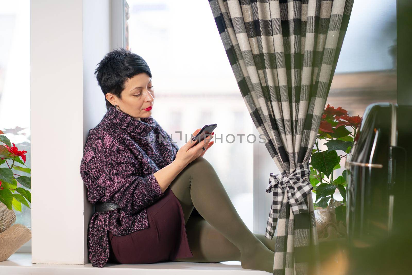 Short hair brunette woman reads news on mobile phone while sitting on windowsill by Try_my_best