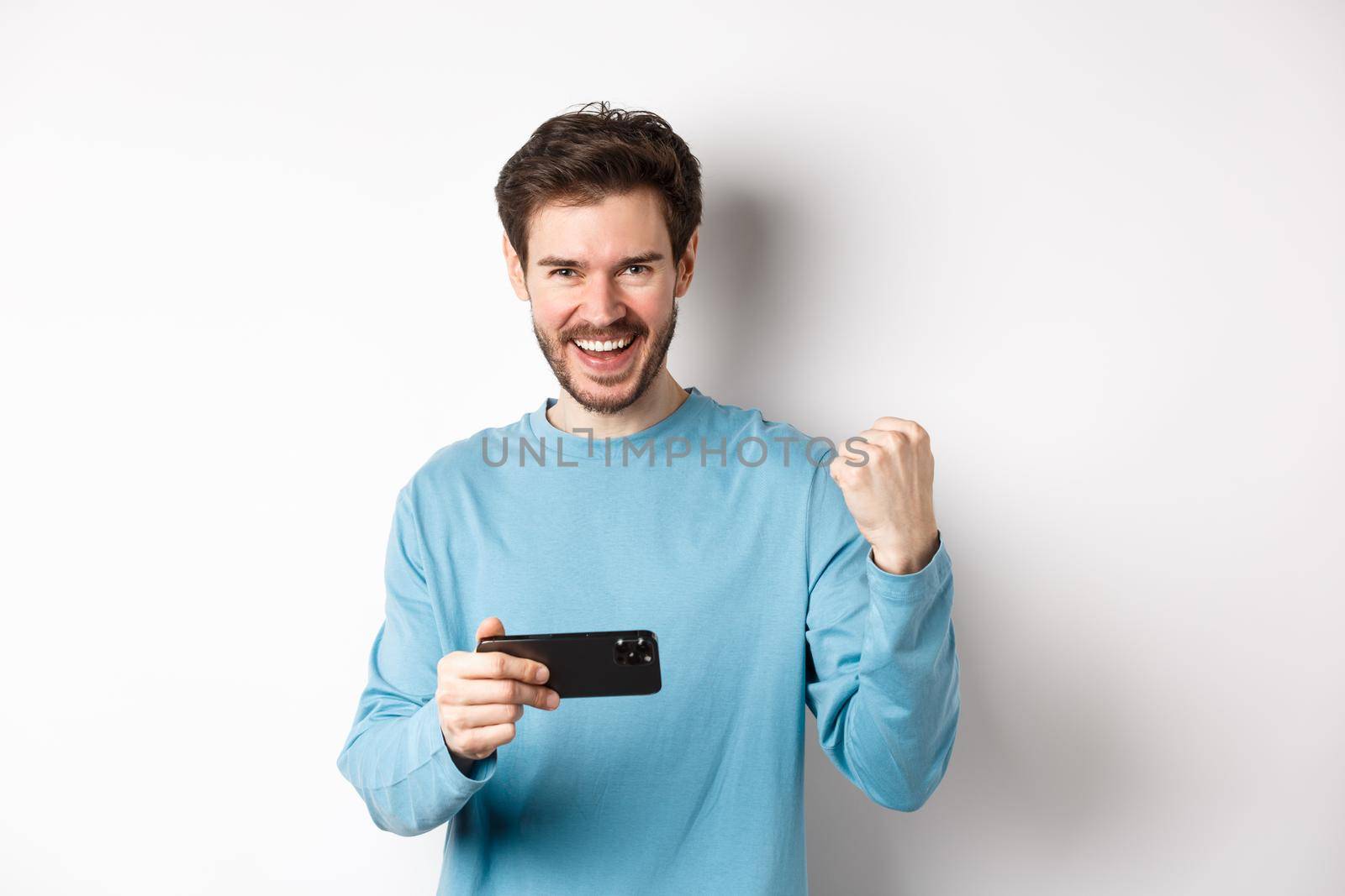 Lucky man winning in mobile video game, holding smartphone and saying yes, triumphing with fist pump, standing over white background.