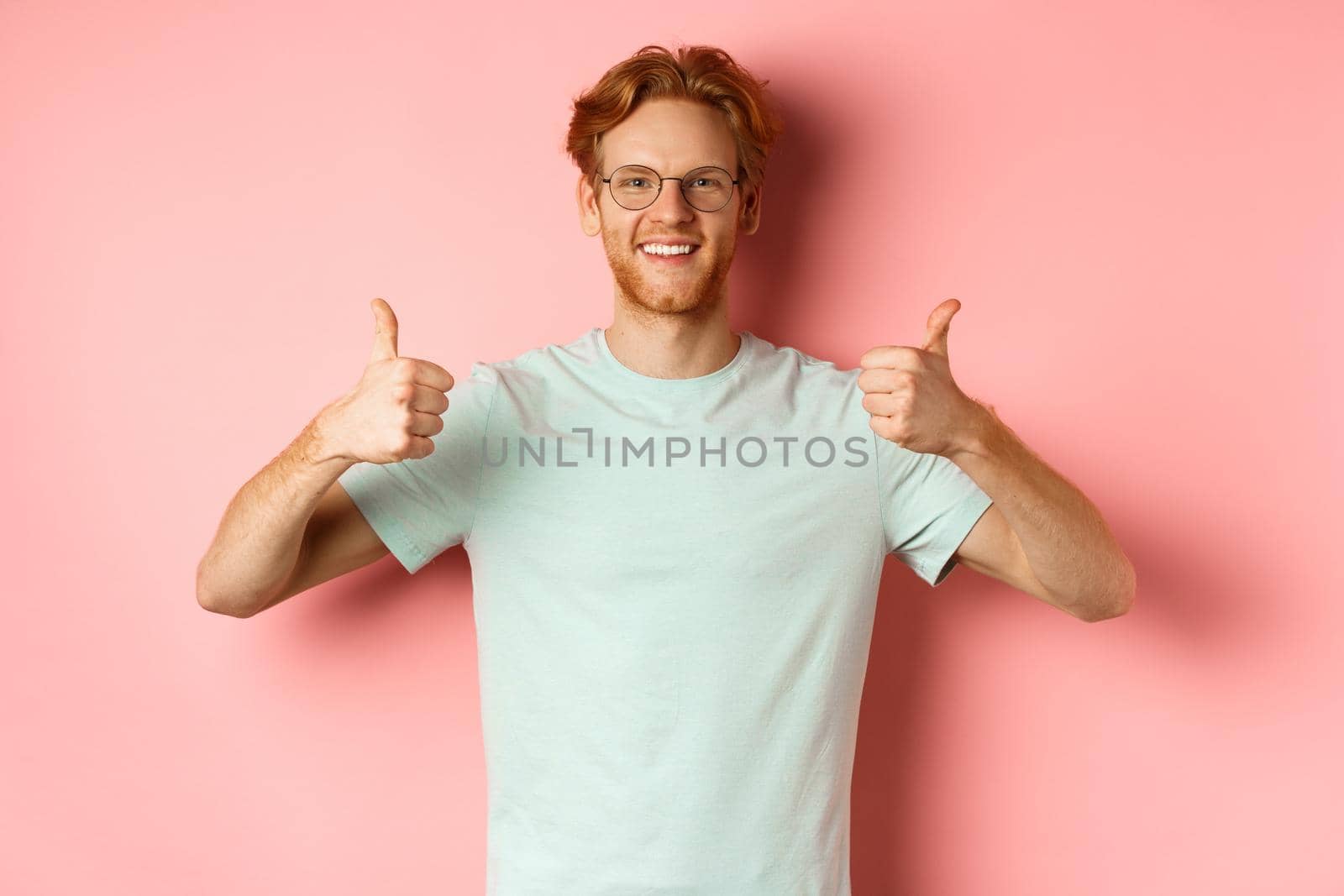 Cheerful european man with red hair and beard, wearing glasses, showing thumbs-up and smiling in approval, praise something good, standing over pink background.