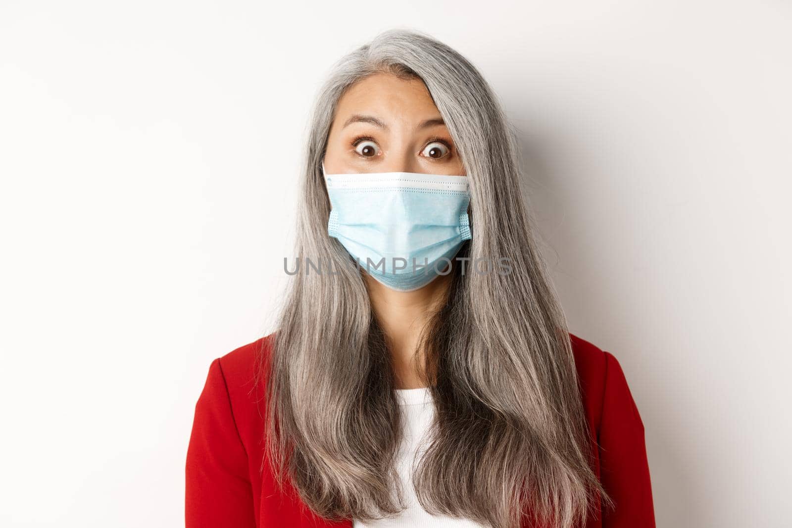 Covid, pandemic and business concept. Close up of surprised asian grandmother in medical mask raising eyebrows, staring amazed at camera, white background.