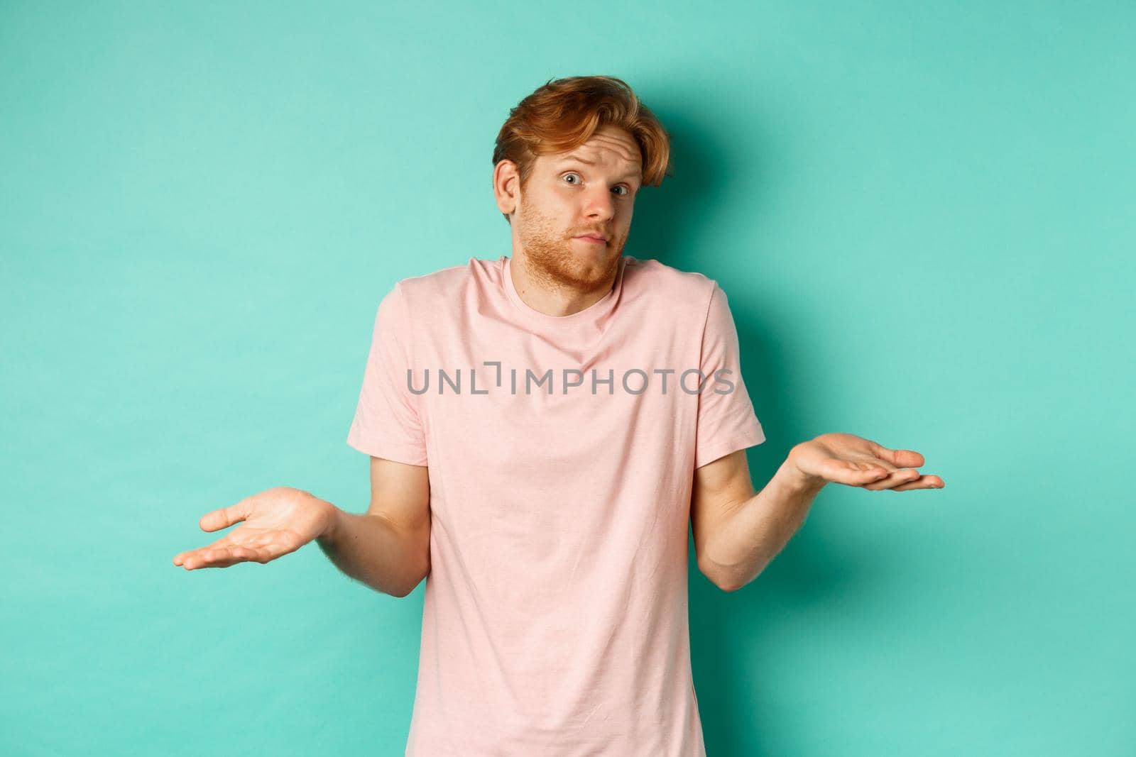 Young redhead guy in t-shirt know nothing, shrugging shoudlers with hands spread sideways, standing confused over turquoise background.