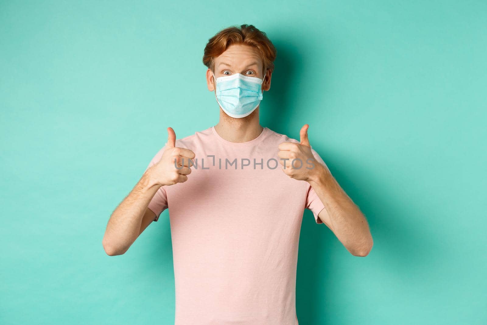 Covid-19, pandemic and lifestyle concept. Cheerful redhead guy in medical mask showing thumbs up in approval, like and praise product, standing over turquoise background.