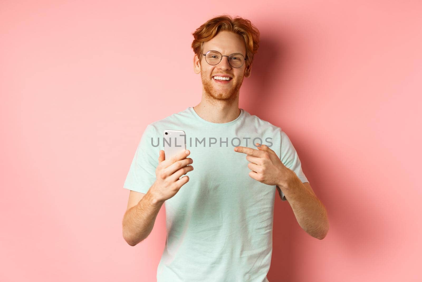 Young man with red hair and beard, wearing t-shirt and glasses, smiling while pointing finger at smartphone, recommend online promotion, pink background by Benzoix