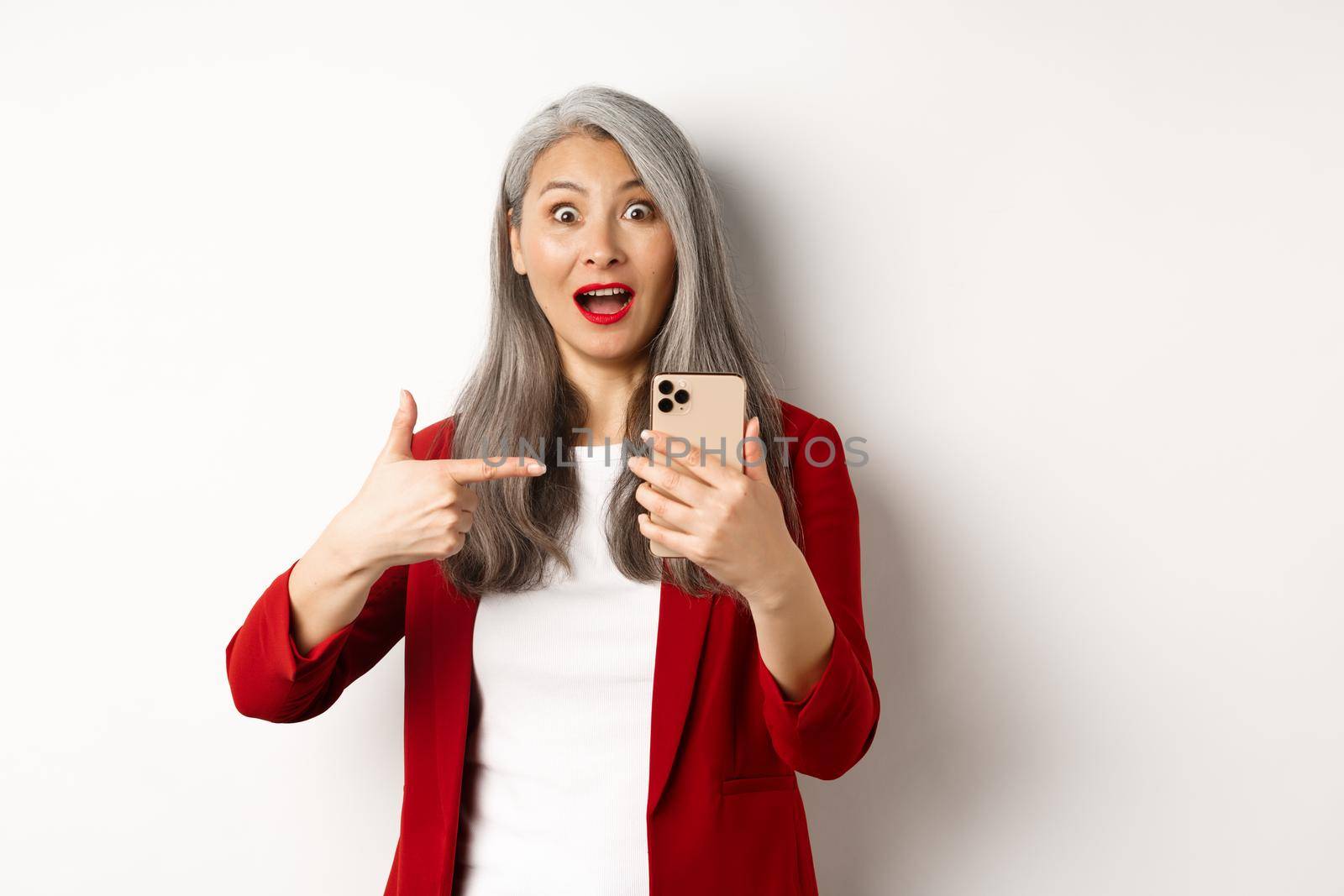 Amazed asian lady in red blazer pointing finger at smartphone, staring impressed at camera, showing something online, white background by Benzoix