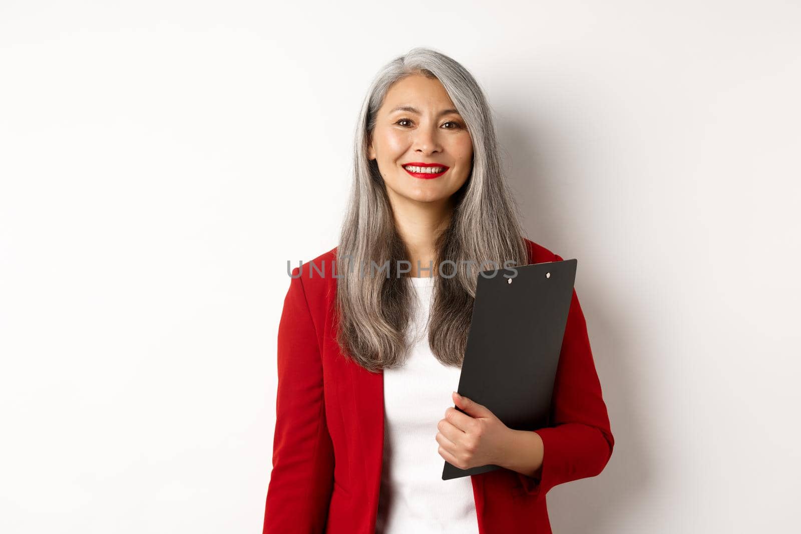 Successful asian senior business woman holding clipboard, wearing red blazer and lipstick at work, smiling at camera, white background by Benzoix