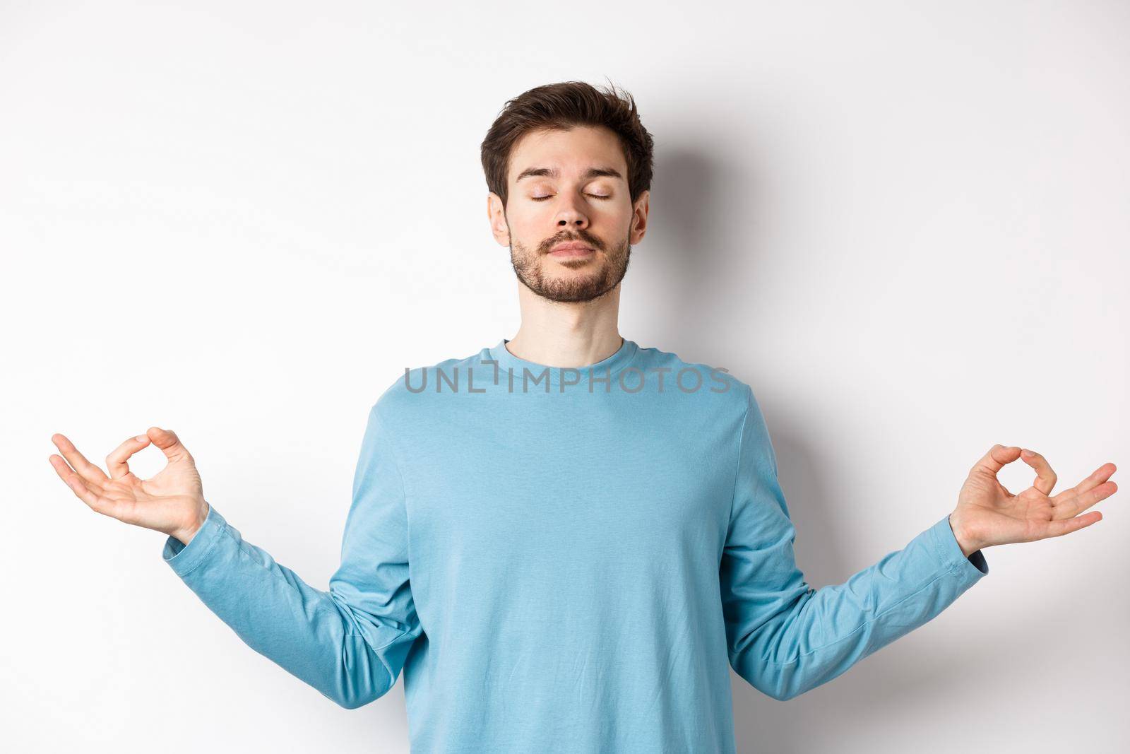Young man trying to relax in meditation, standing calm with stretch out hands in zen mudra gesture, practice yoga on white background.