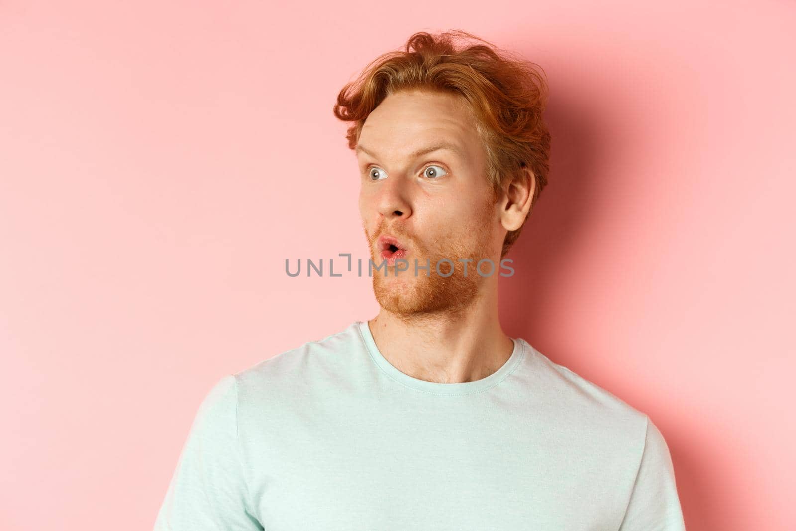 Headshot portrait of surprised redhead man with beard, looking left and saying wow, raising eyebrows amazed, standing over pink background.
