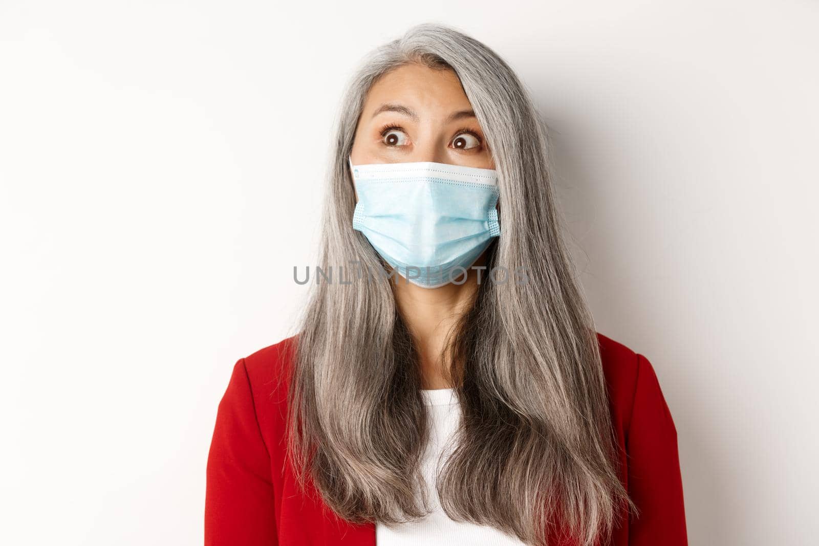 Covid, pandemic and business concept. Close up of elegant asian senior lady staring at upper left corner, wearing medical mask from coronavirus, white background by Benzoix