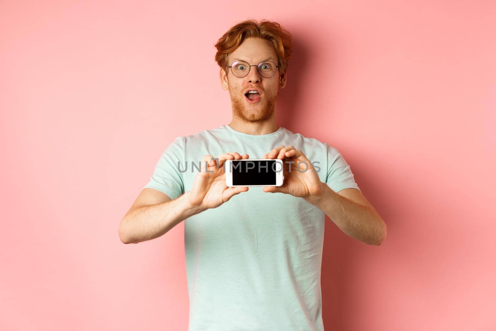 Amazed redhead man gasping and staring with awe at camera, showing blank smartphone screen horizontally, standing over pink background.