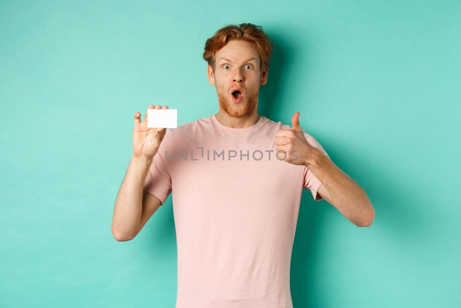 Adult guy with red hair and beard showing plastic credit card and thumb up, looking impressed, recommend bank, standing over mint background.