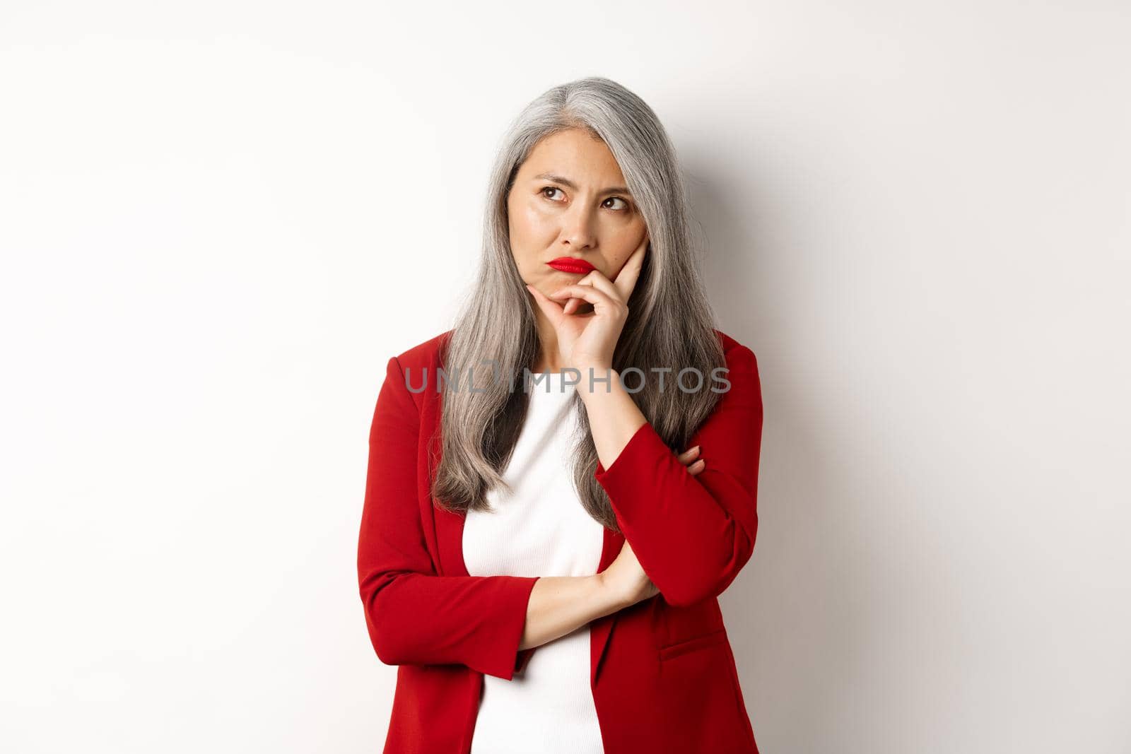 Troubled asian businesswoman in red blazer and lips, pouting and looking left annoyed, standing against white background.