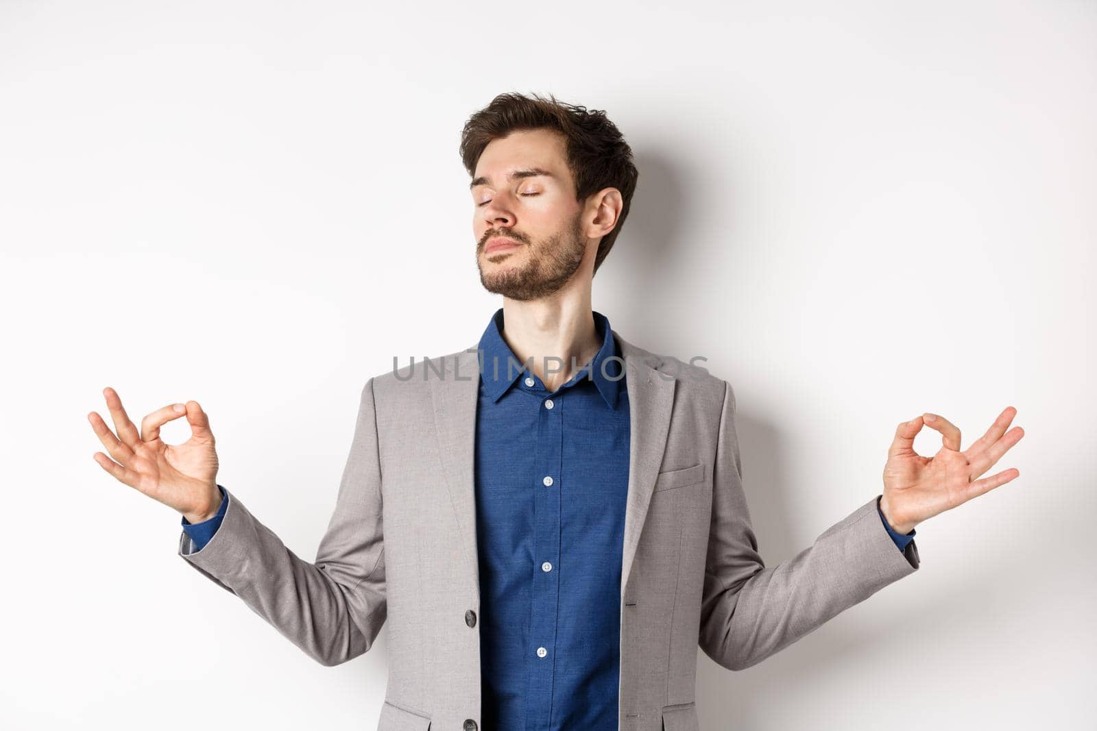 Calm and focused businessman meditating with eyes closed and hands spread sideways, finding peace in meditation, practice yoga breathing, standing on white background.