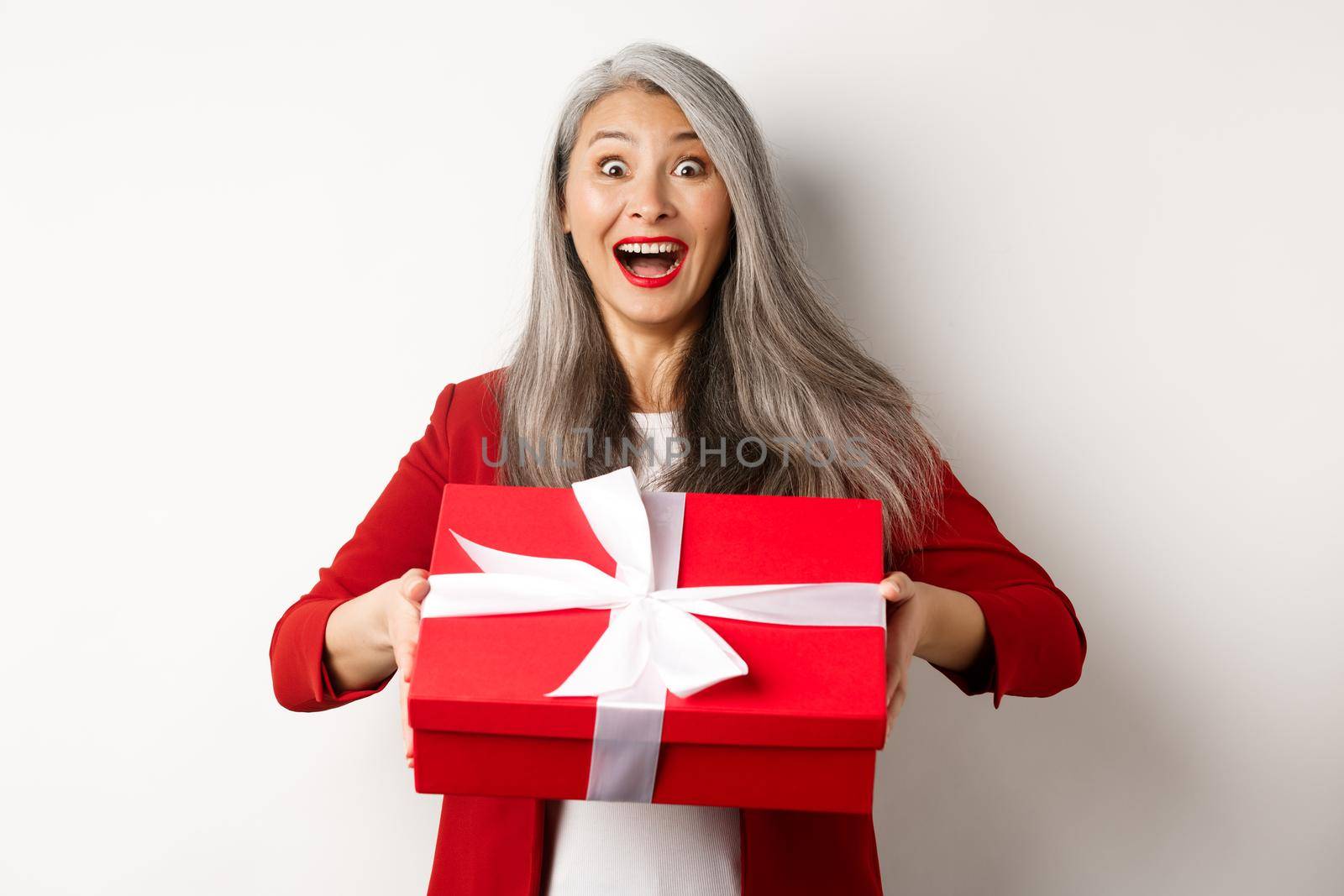 Surprised and thankful asian grandmother receiving gift in red box, looking amazed and grateful, standing over white background by Benzoix