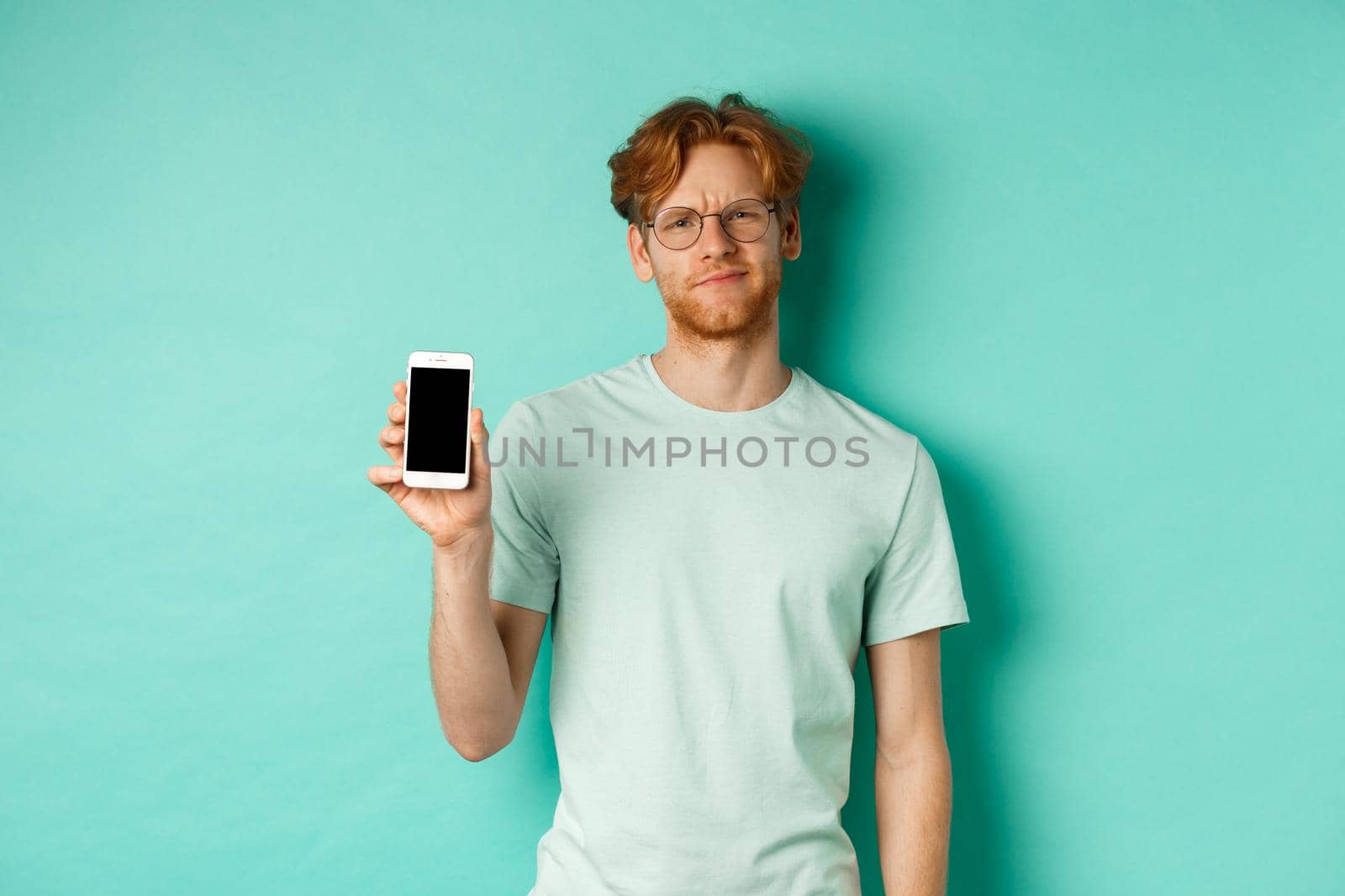 Skeptical male model with red hair and glasses, showing mobile screen and frowning disappointed, dislike application, standing over turquoise background.