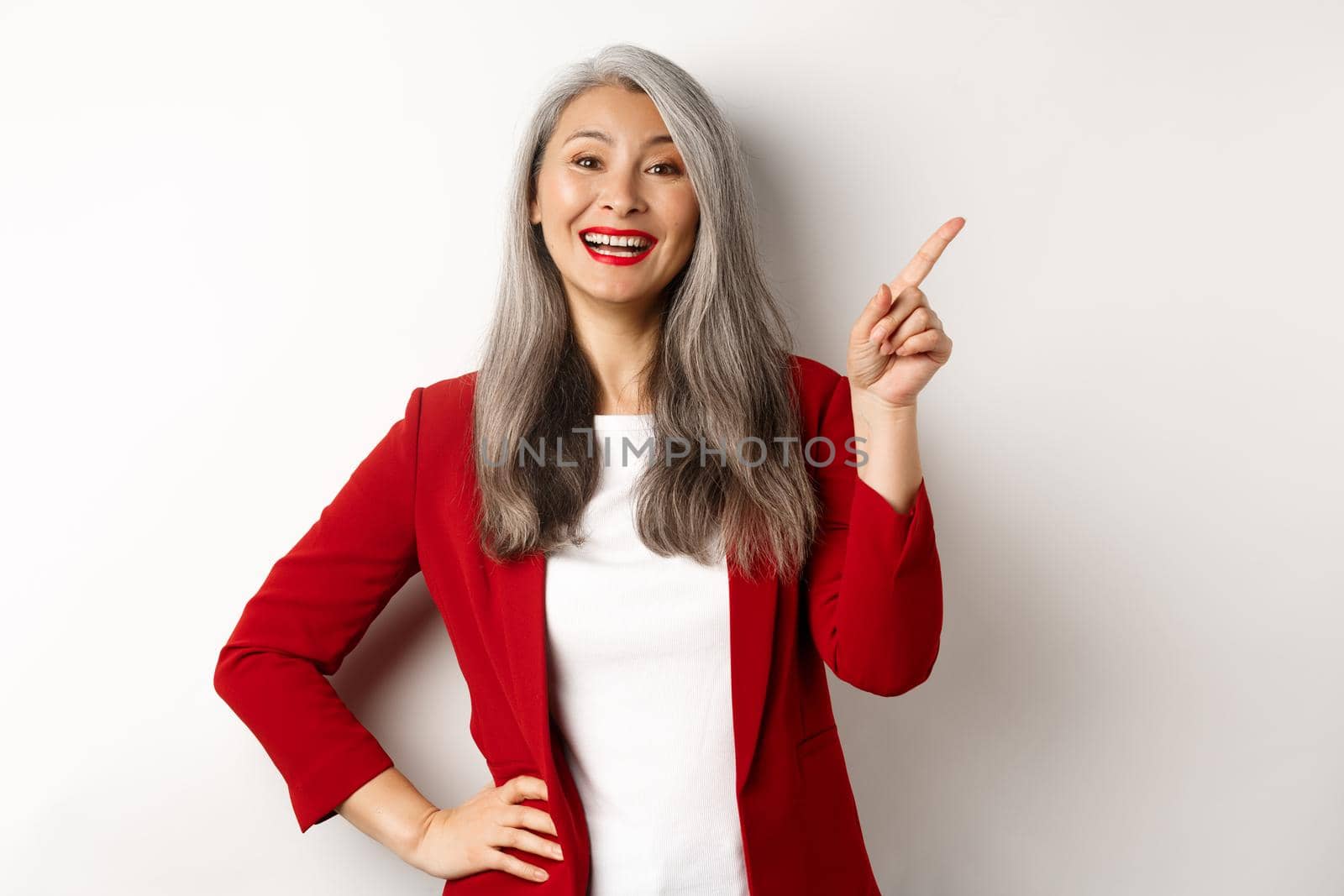 Asian professional businesswoman showing logo, smiling and pointing finger upper right corner, standing over white background.