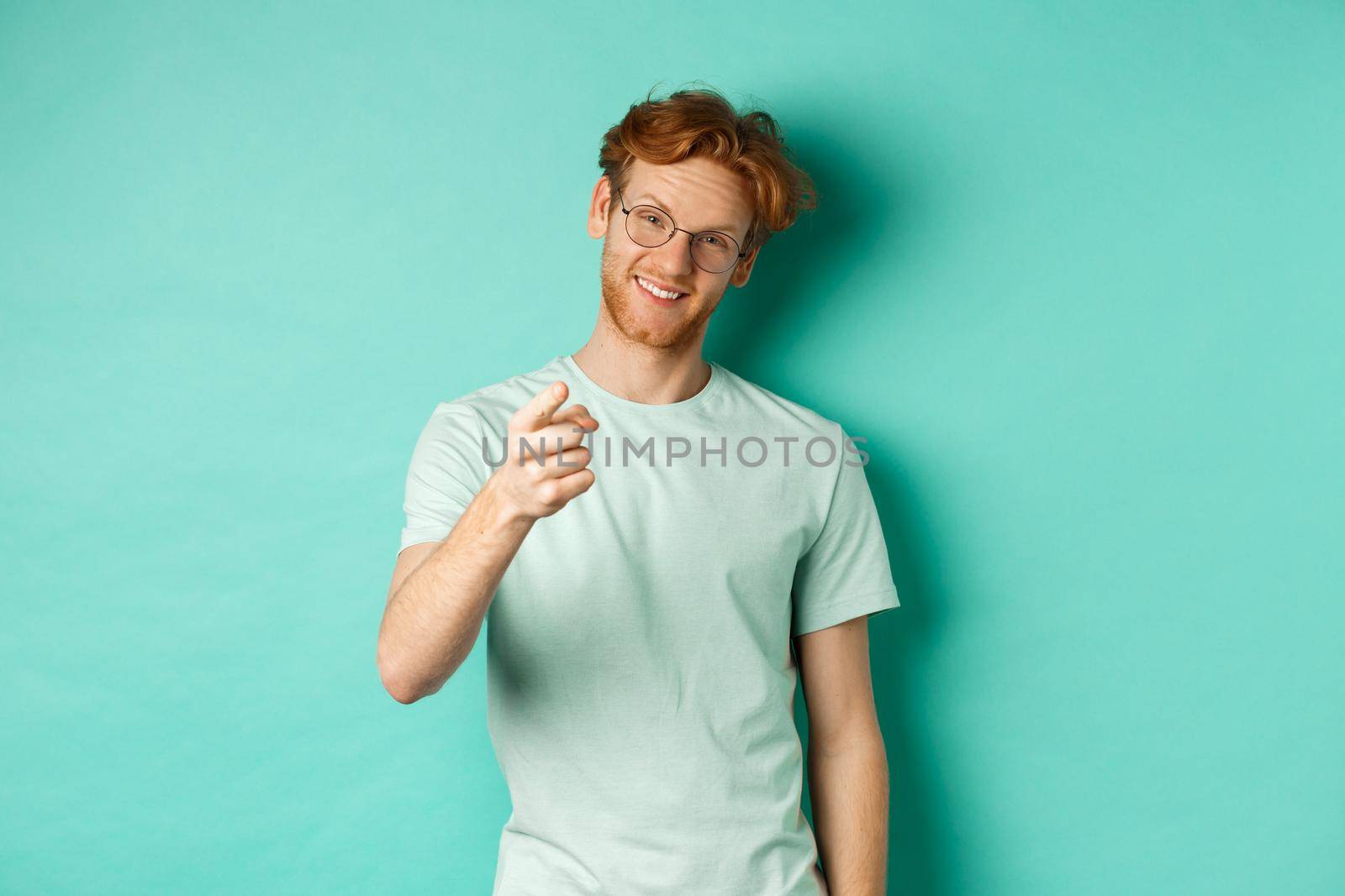 Handsome young man with ginger hair, wearing glasses and t-shirt, pointing finger at camera and smiling, choosing you, making invitation, standing over turquoise background.
