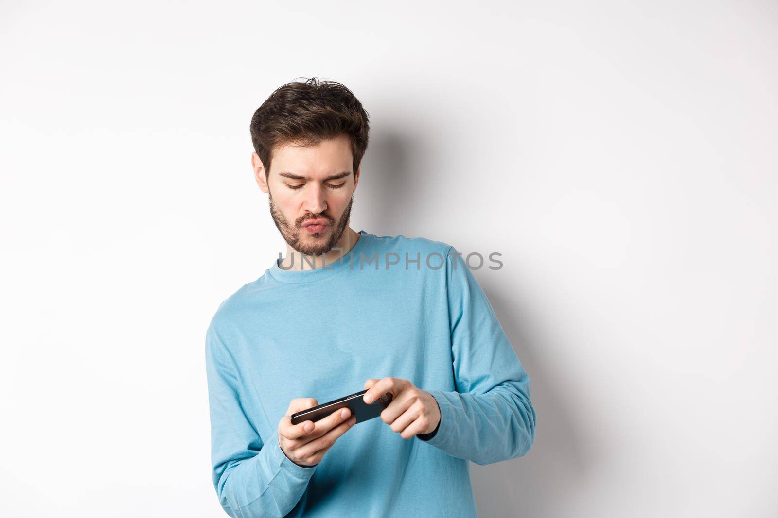 Young man playing video games on smartphone, tilt body and tap mobile screen, standing joyful over white background by Benzoix