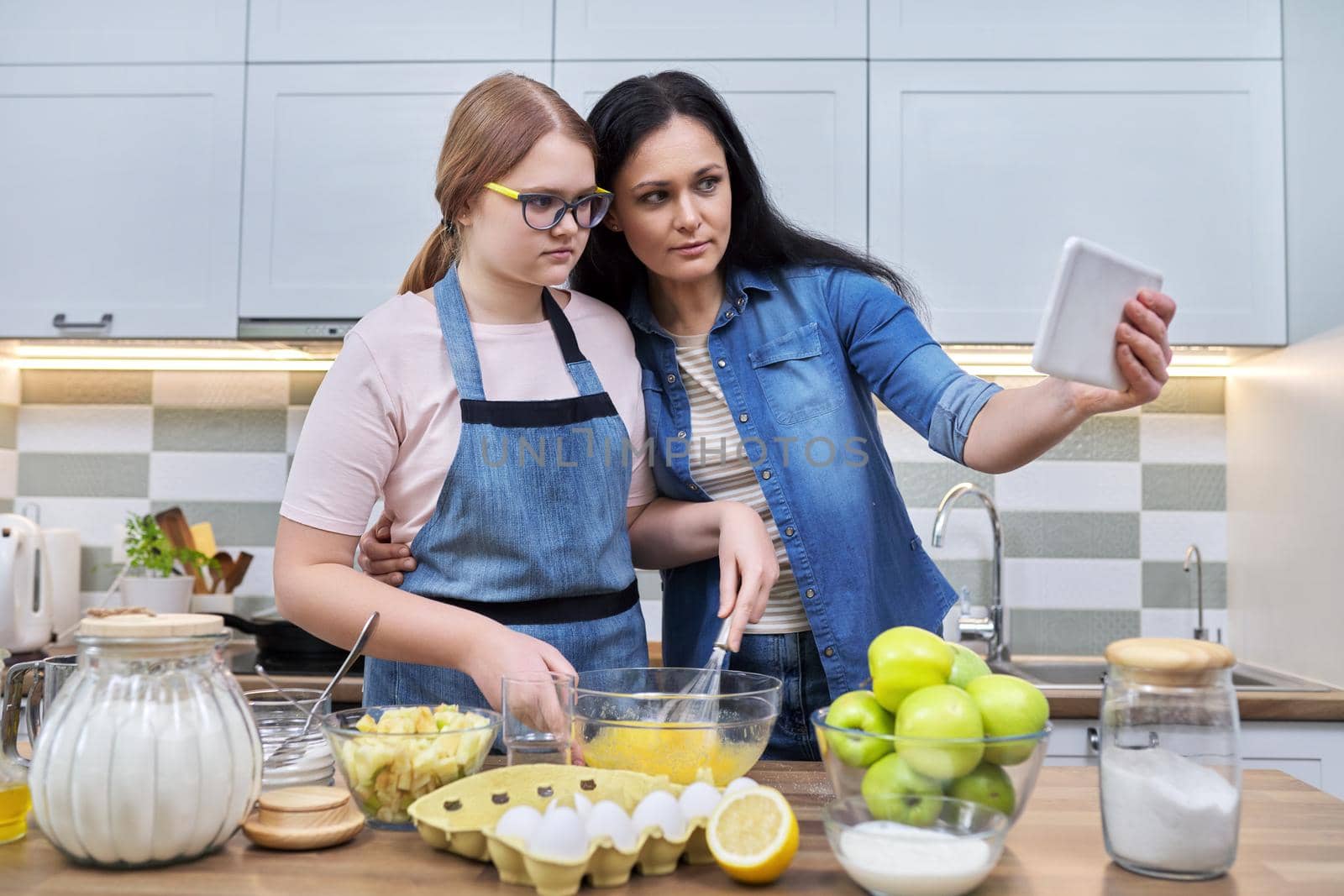 Mom and teen daughter preparing apple pie together, at home in kitchen. Woman with digital tablet, recipe, video call, technology in daily life. Family, parent teenager relationship, lifestyle