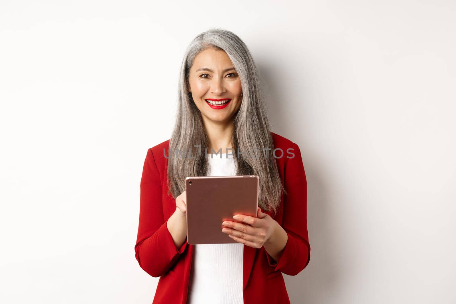 Business. Senior female entrepreneur working on digital tablet and smiling happy at camera, wearing red blazer and makeup, standing over white background.