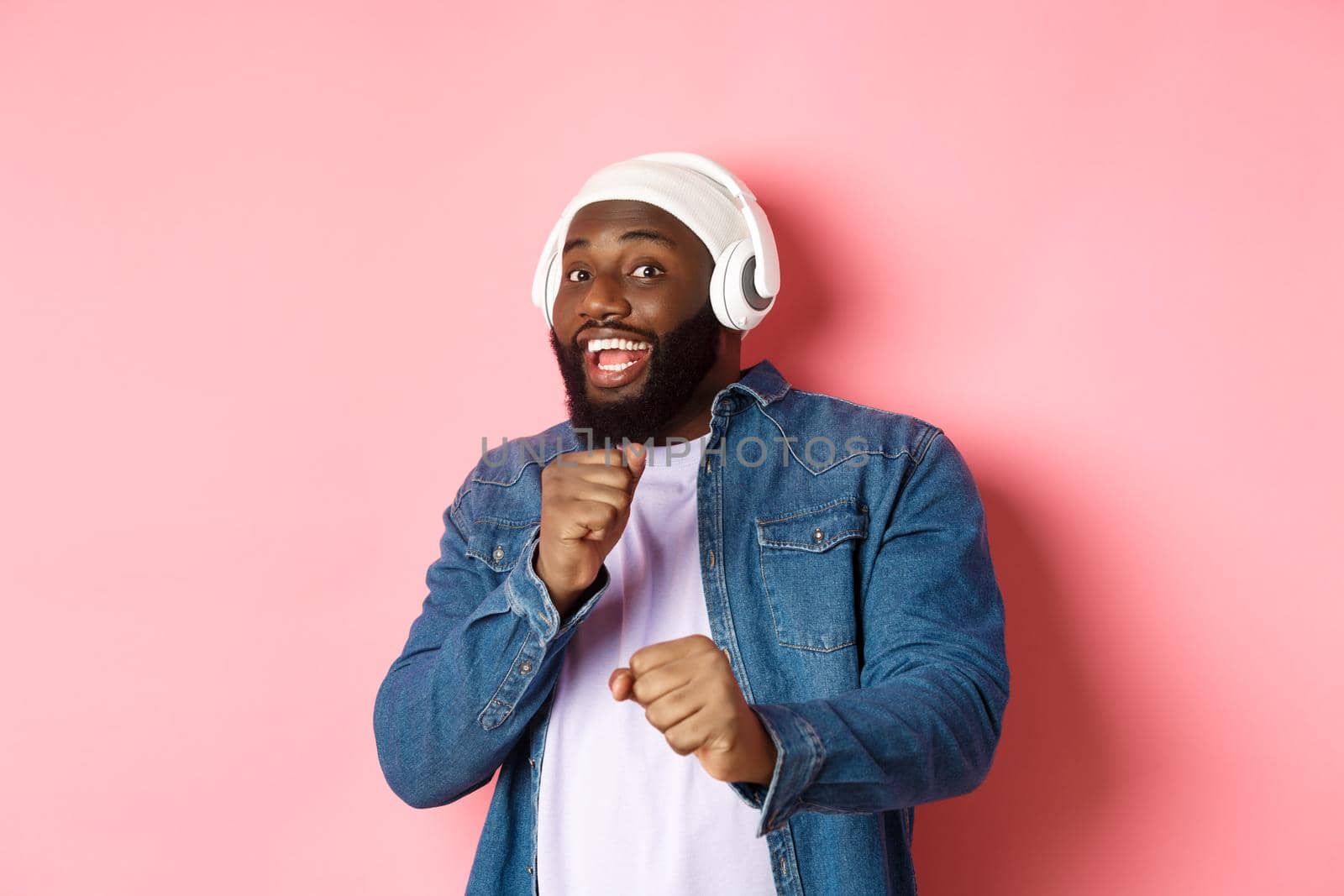 Happy african-american man dancing, listening music in headphones and looking at camera, standing over pink background.