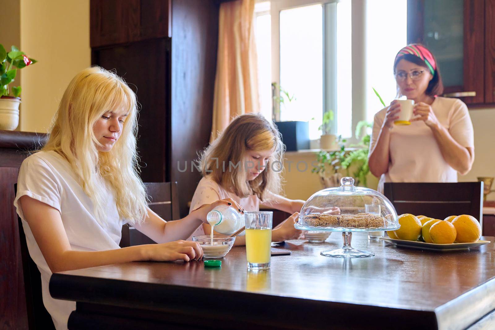 Family, girls children eating at table in kitchen. Home kitchen interior, mother with a cup, sisters eating and looking at smartphone screen. Lifestyle, daily routine, home, people, technology