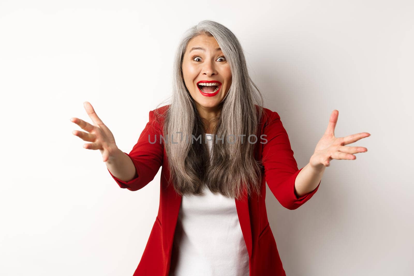 Surprised and friendly asian woman reaching hands forward and smiling, welcoming you, greeting someone, standing over white background.