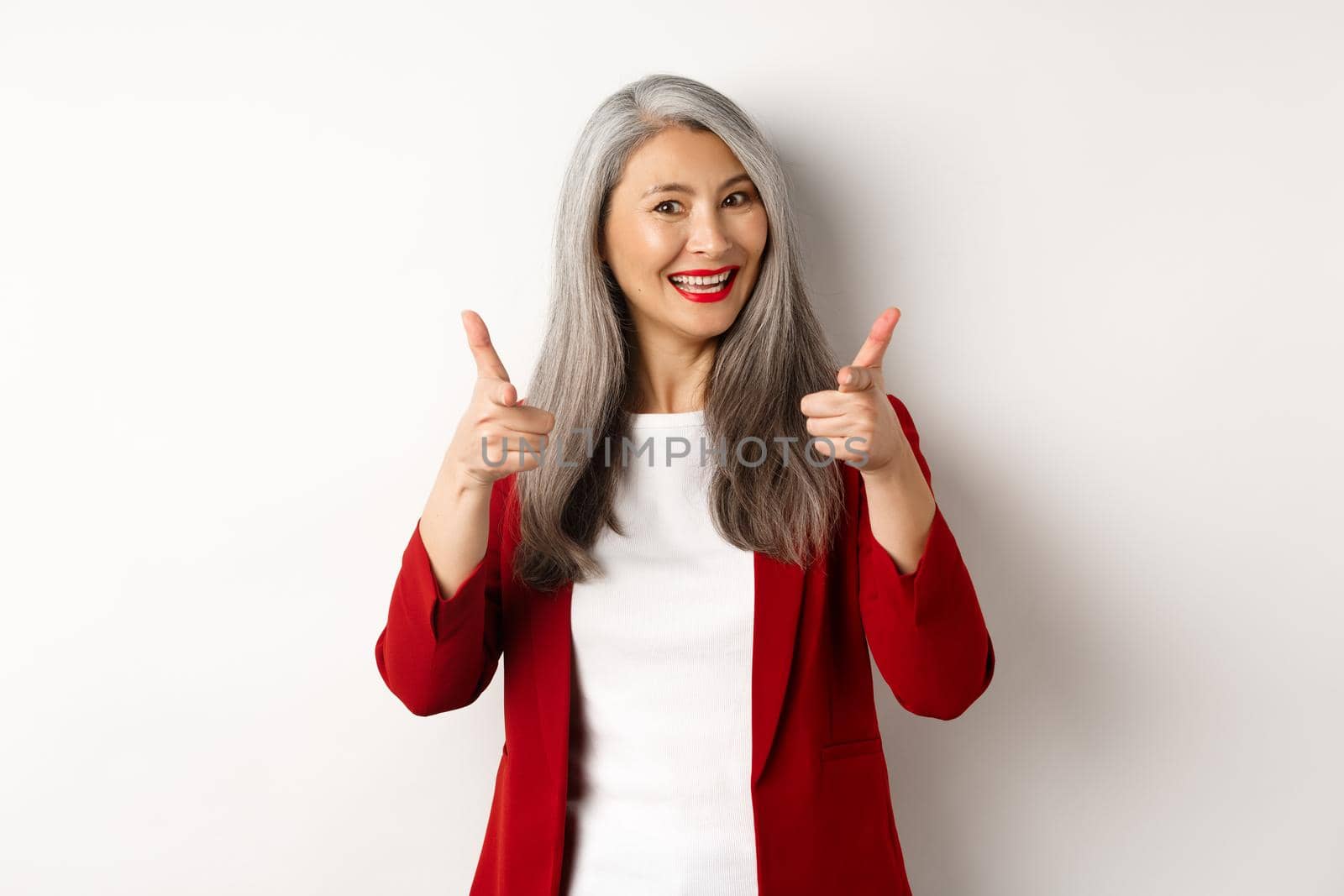 Professional female employer in trendy red blazer and makeup, pointing fingers at camera and smiling, praising something, need you, standing over white background.