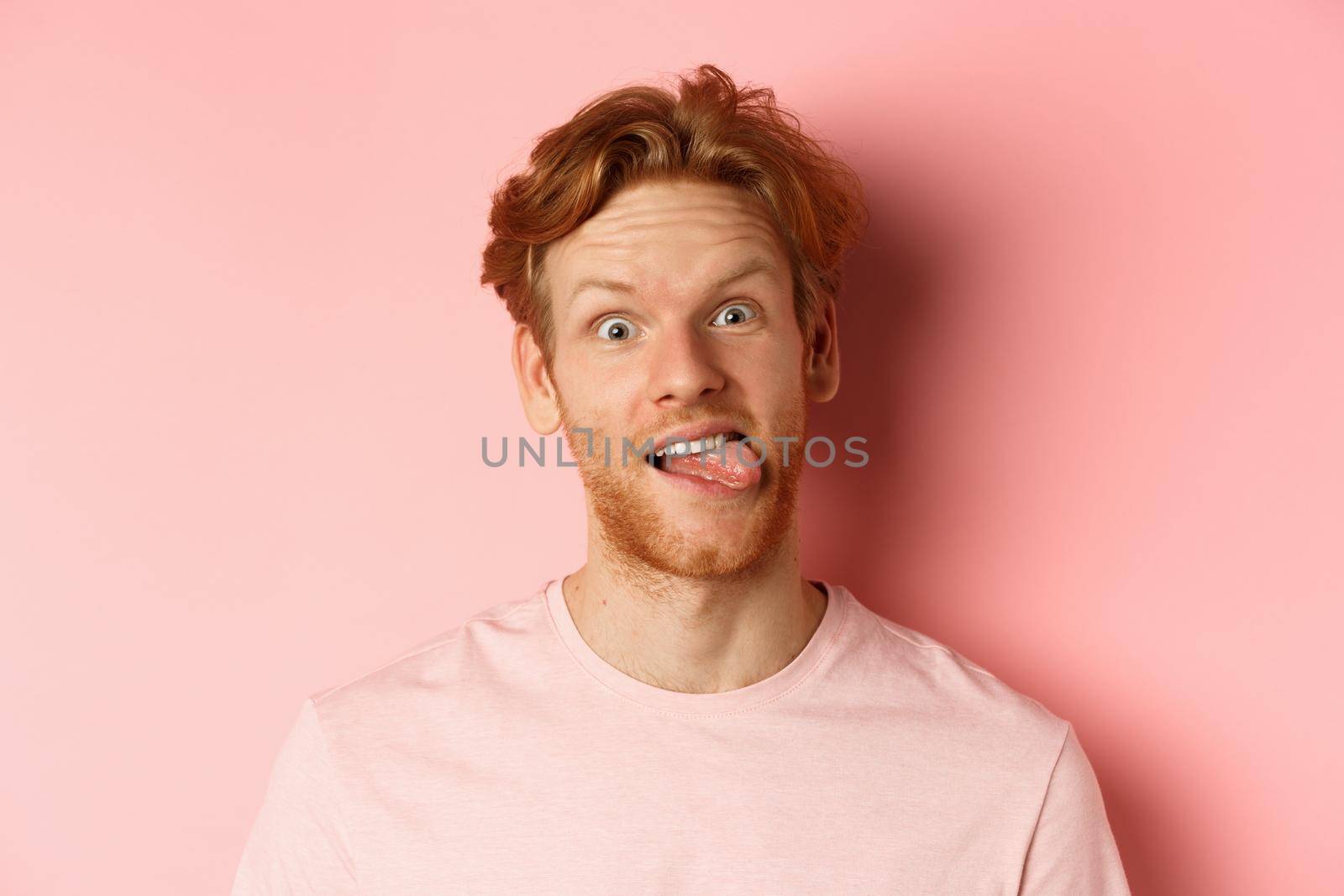Headshot of funny redhead guy showing tongue, making silly faces at camera, standing joyful against pink background.