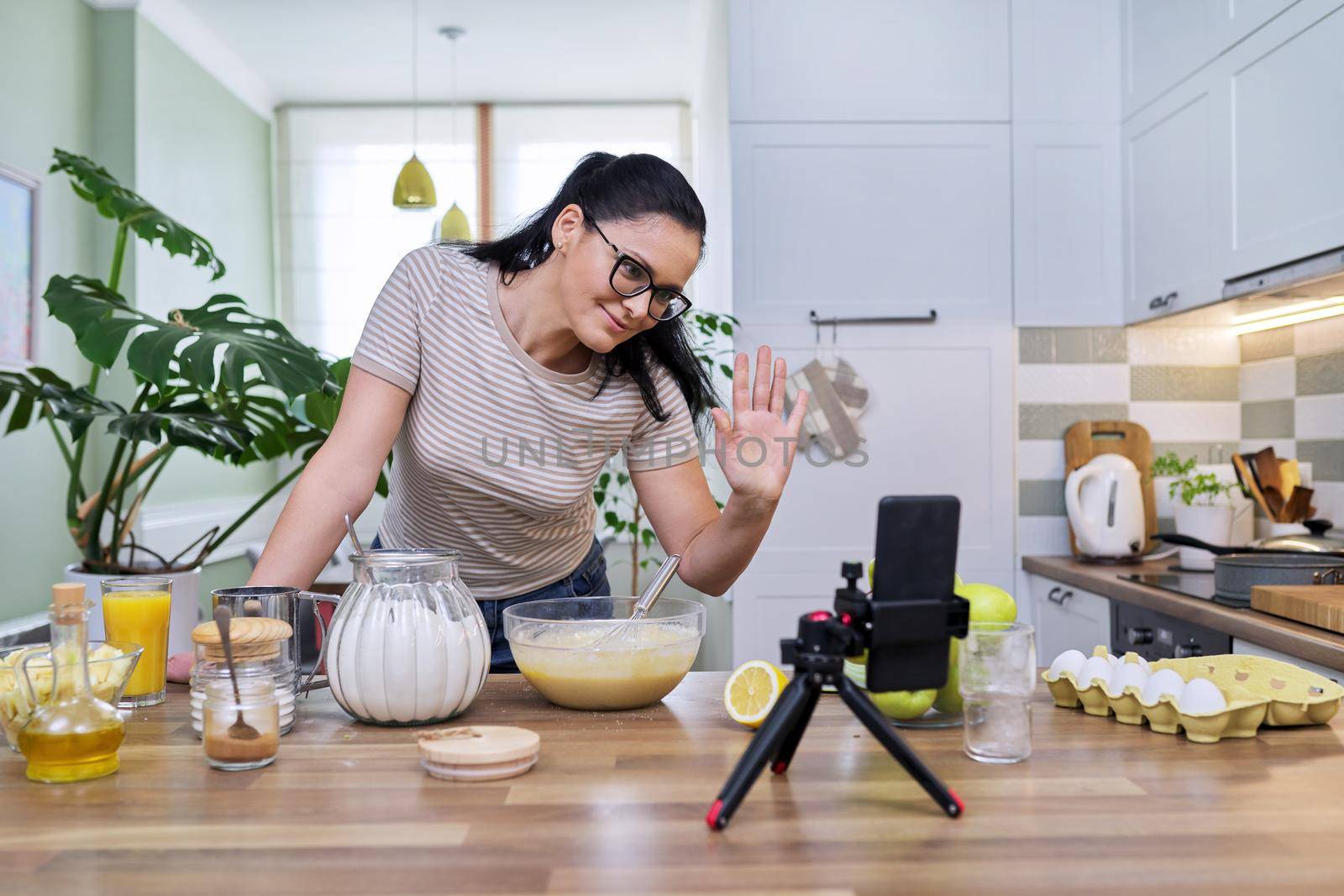 Technologies in everyday life, 40s woman preparing apple pie at home in kitchen, with smartphone on tripod using video call for communication. Lifestyle, eating at home, technology, people concept