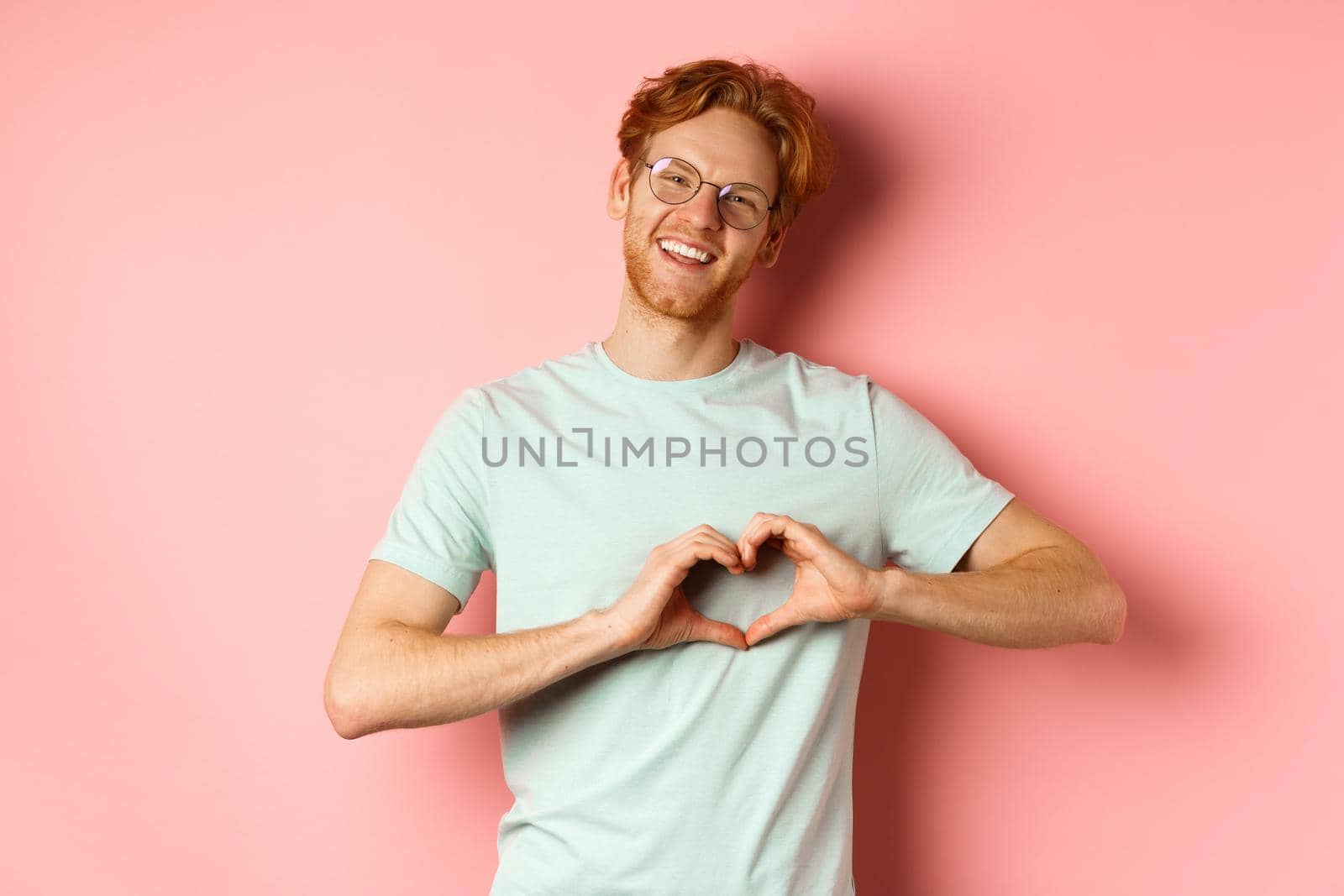Valentines day concept. Handsome redhead man in glasses, showing heart sign and say I love you, standing over pink background.