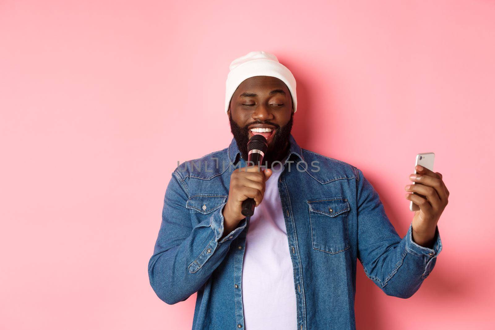 Handsome african-american man singing karaoke, reading lyrics on smartphone app and holding microphone, standing over pink background.