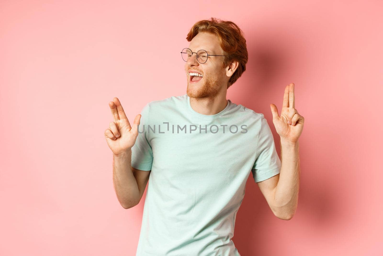 Cheerful redhead guy listening music and dancing, triumphing or celebrating success, standing over pink background.