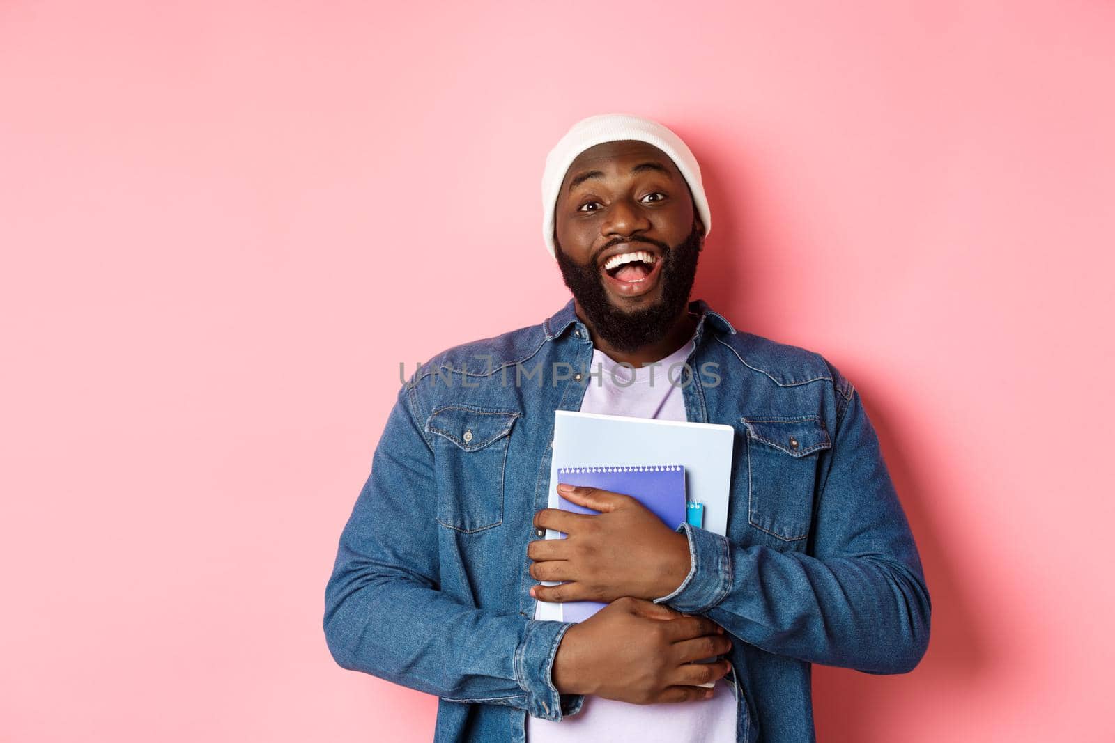 Education. Happy african-american male student in beanie holding notebooks, studying courses, smiling at camera, standing over pink background by Benzoix