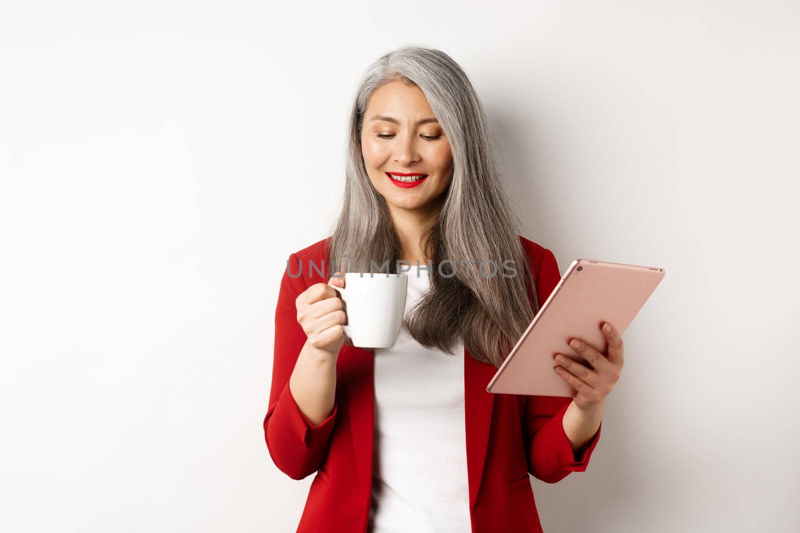 Business people concept. Asian mature businesswoman holding digital tablet and looking at coffee cup with pleased smile, standing over white background.