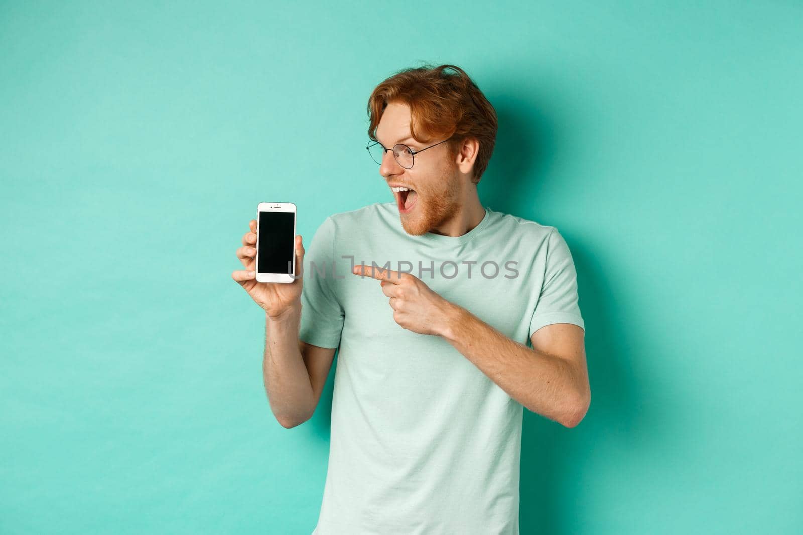 Check this out. Handsome redhead guy in glasses pointing finger at blank smartphone screen, showing online promotion, standing amazed over turquoise background.