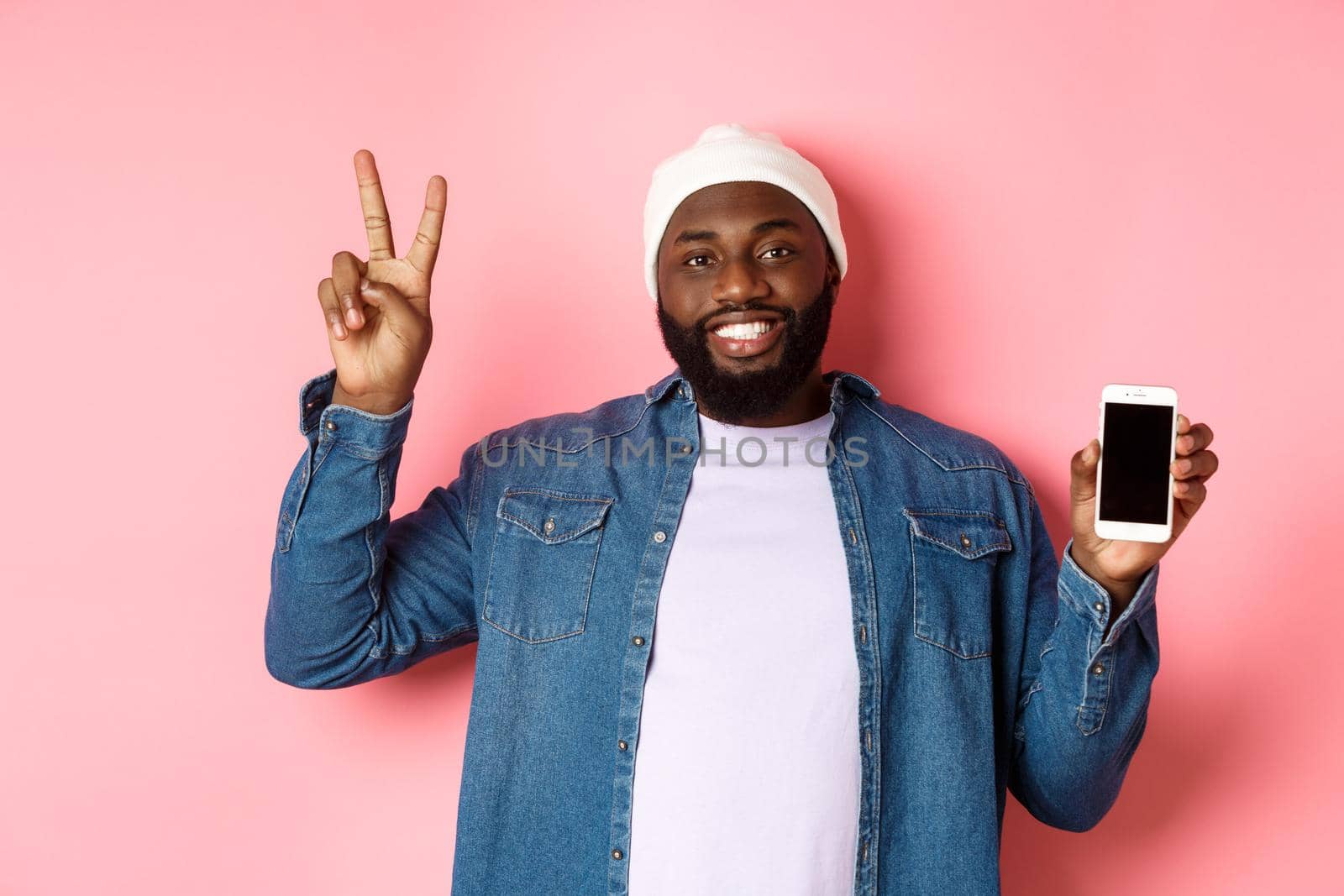Online shopping and technology concept. Happy Black guy in beanie and denim shirt showing mobile screen and peace sign, standing over pink background.