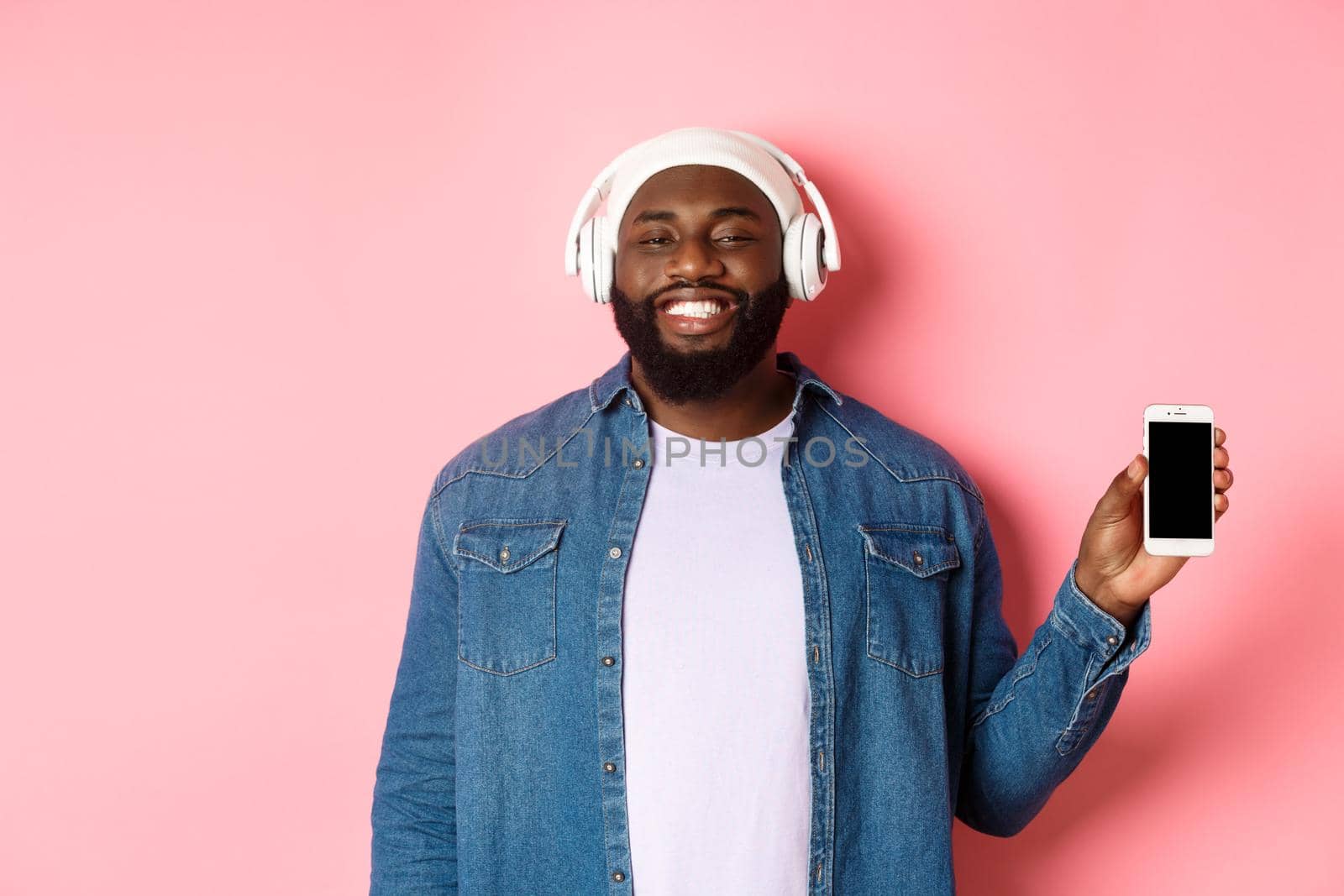 Happy hipster guy listening music on headphones and showing mobile screen, smiling satisfied, standing over pink background.