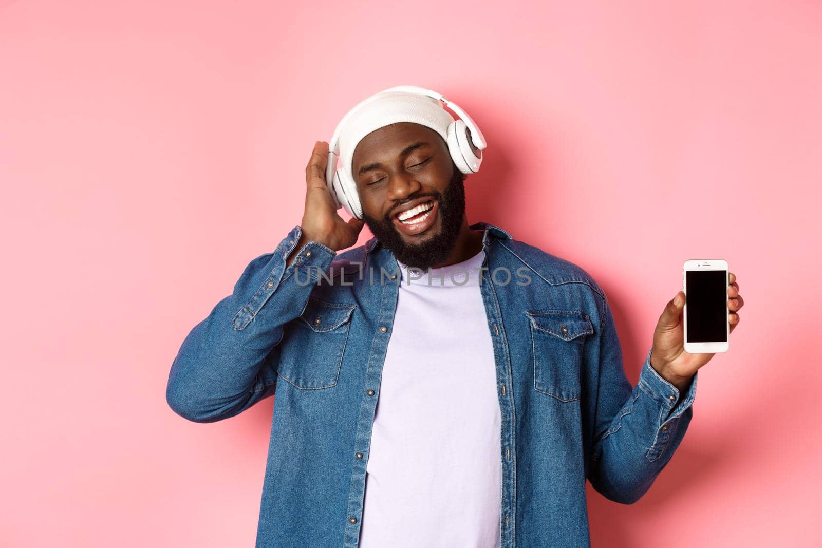 Carefree handsome Black man listening music in headphones, enjoying sound and showing smartphone screen, standing with pleased smile on face against pink background.