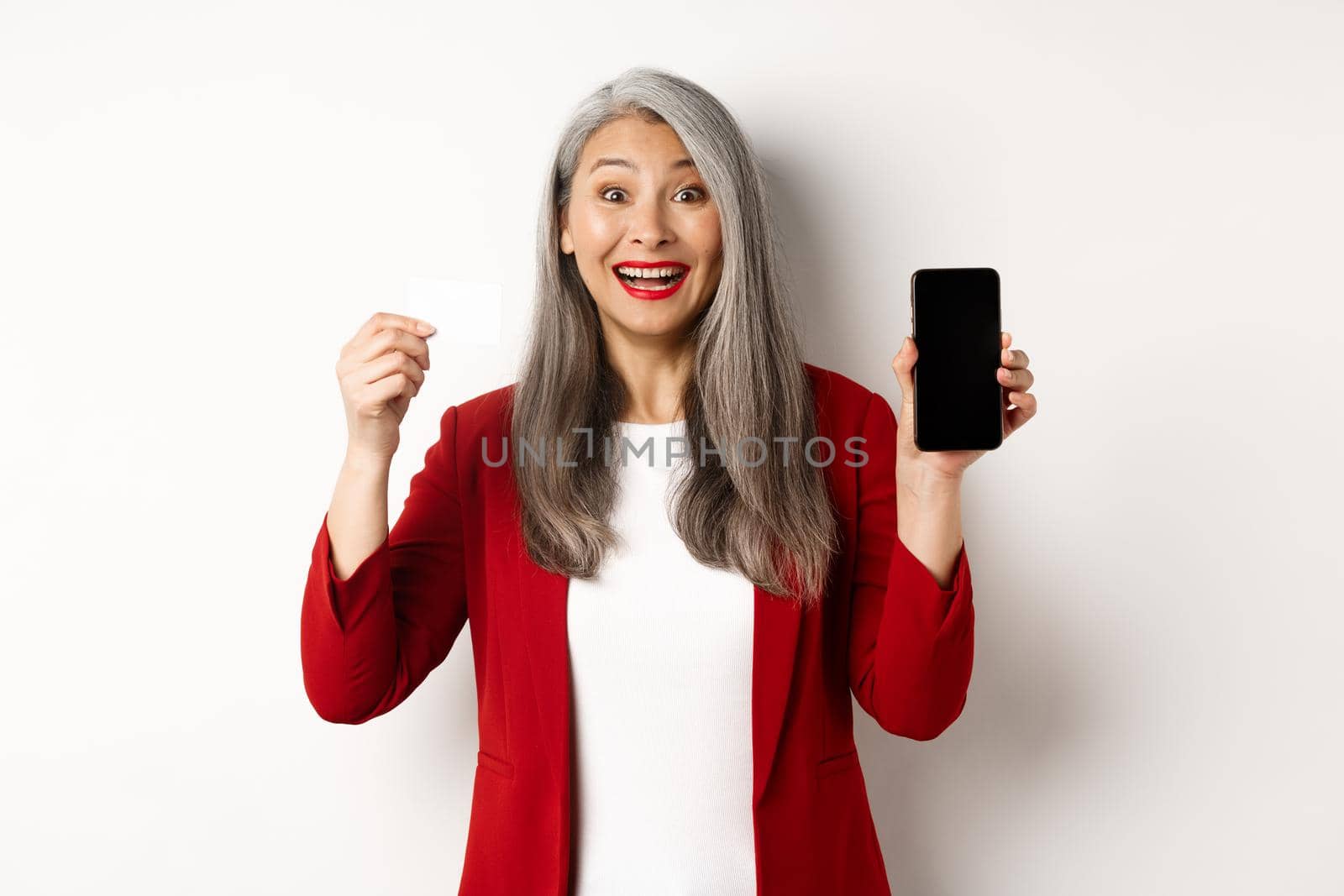 Cheerful asian mature woman showing blank smartphone screen and credit card, concept of e-commerce.