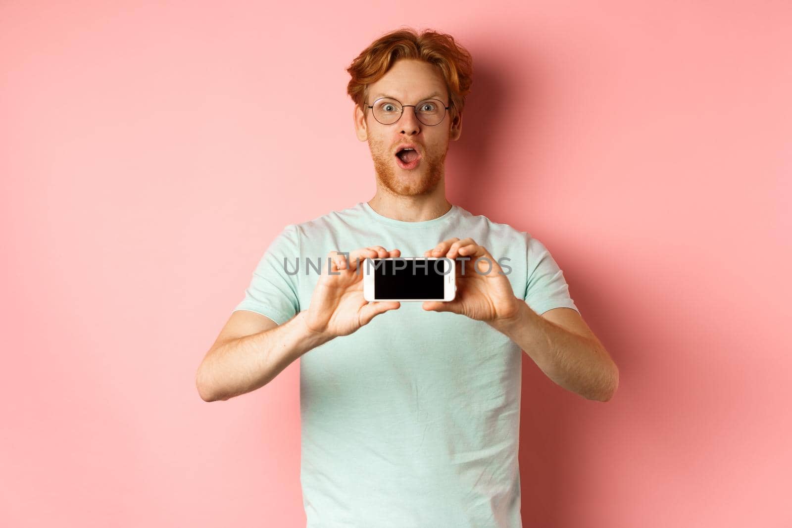 Amazed redhead man gasping and staring with awe at camera, showing blank smartphone screen horizontally, standing over pink background.