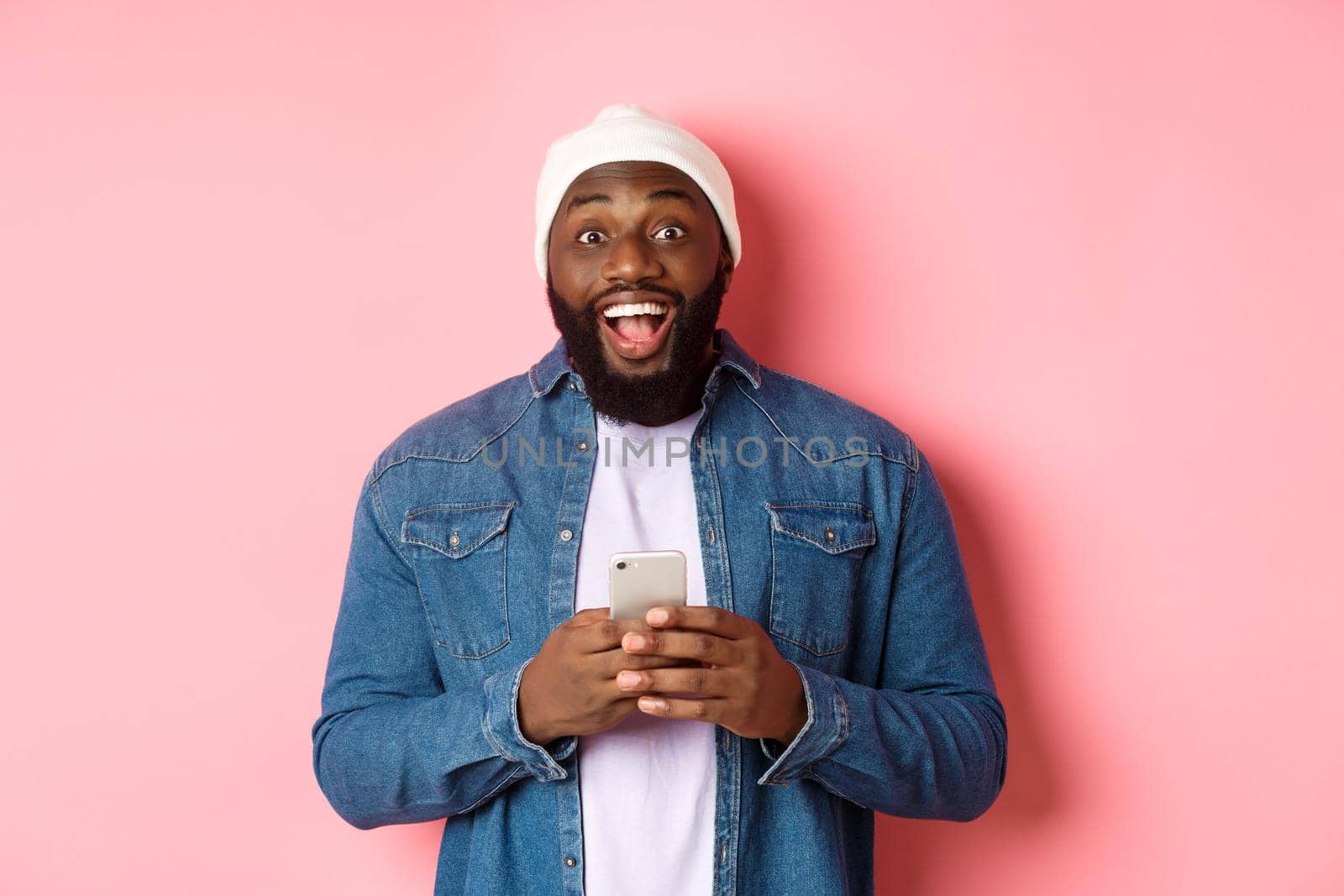 Technology and online shopping concept. Surprised young Black man using mobile phone, looking at camera amazed and happy after reading message, pink background.