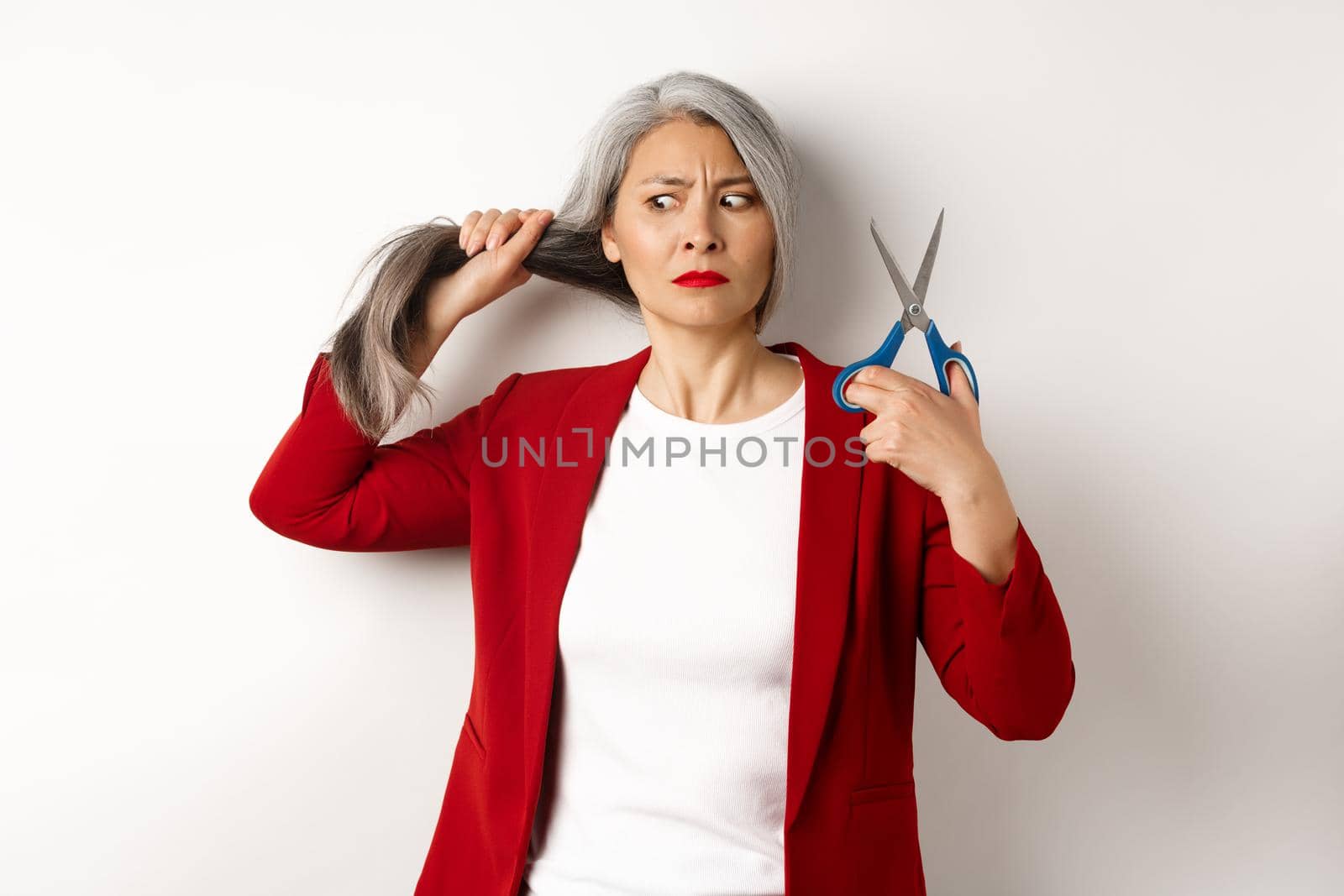 Indecisive asian woman holding scissors and looking doubtful, thinking to cut hair, changing haircut, standing over white background.
