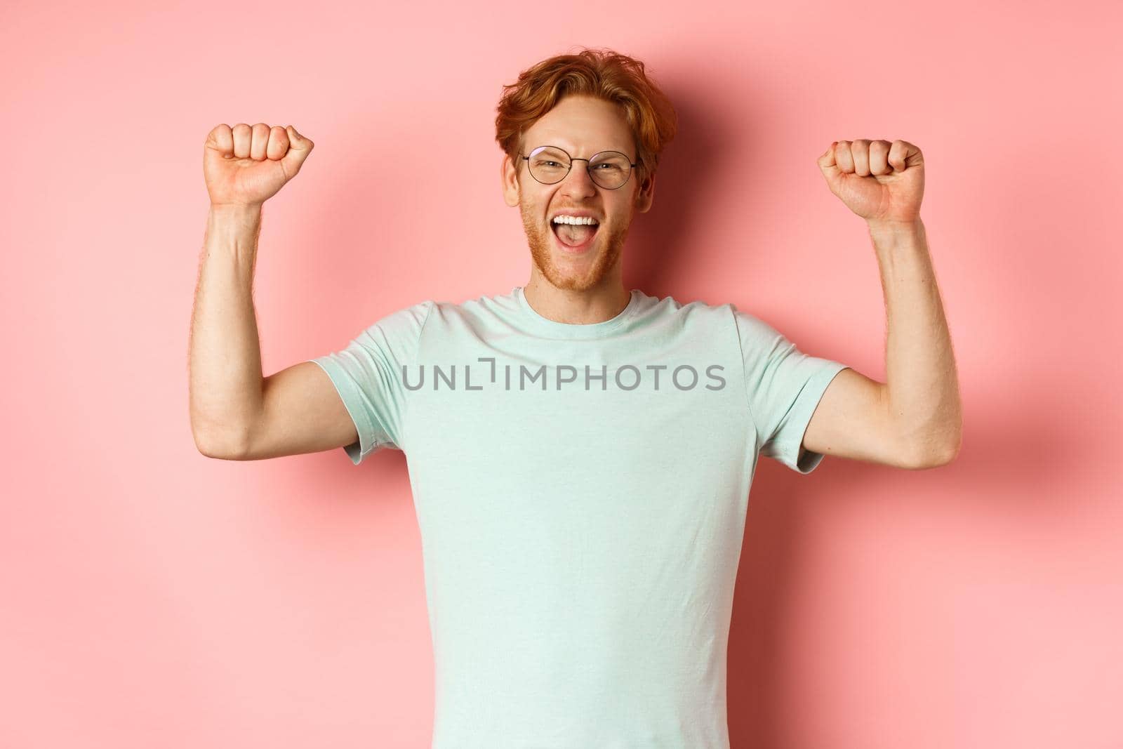Happy winner with red hair and beard celebrating victory, shouting yes with joy and raising hands up, enjoy success, standing over pink background.