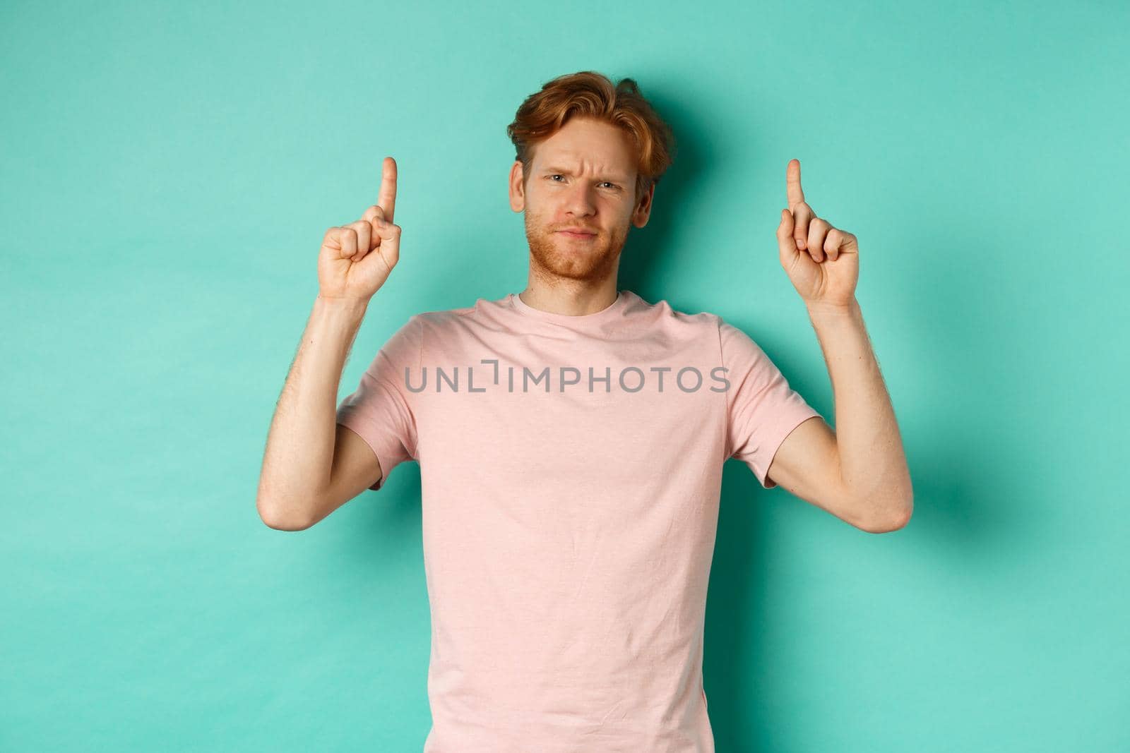 Skeptical and disappointed redhead man pointing fingers up, frowning displeased and looking upset, standing over turquoise background.