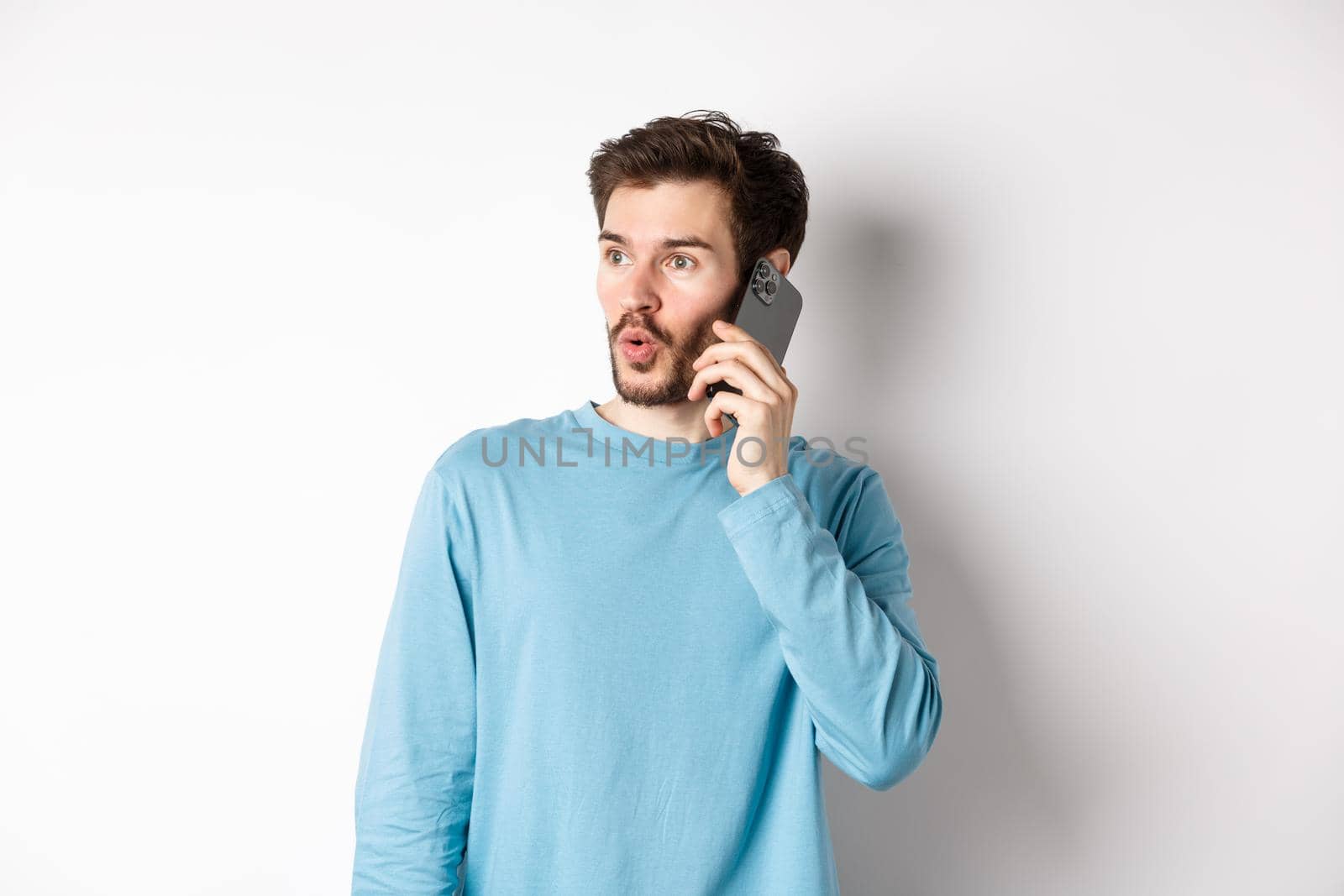 Technology concept. Intrigued young man looking left impressed, talking on mobile phone, calling someone, standing over white background by Benzoix