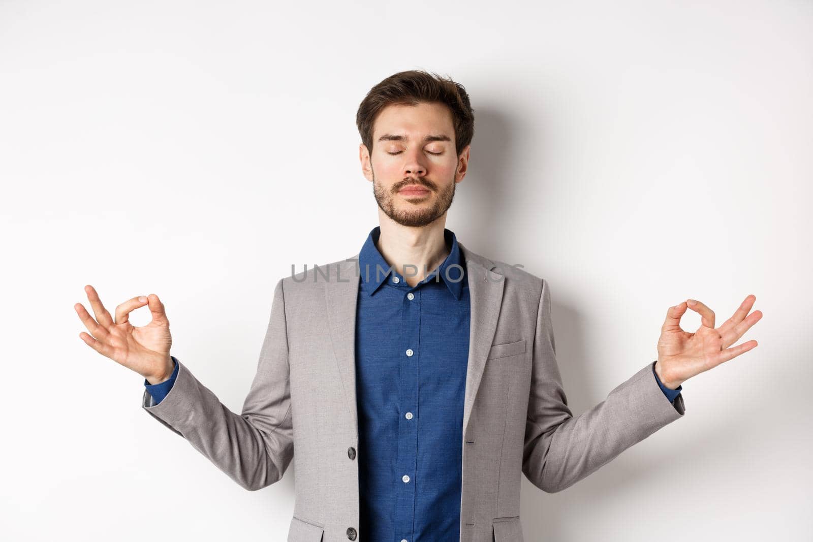 Calm and focused businessman meditating with eyes closed and hands spread sideways, finding peace in meditation, practice yoga breathing, standing on white background.