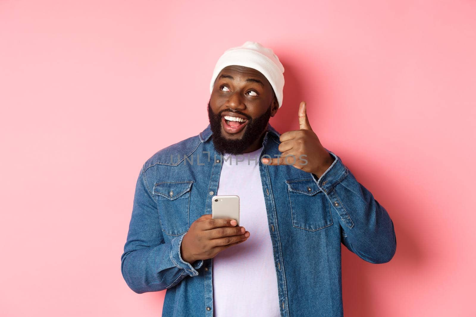 Happy Black man showing call me sign, making phone gesture and smiling, holding smartphone, standing in beanie and denim shirt over pink background.